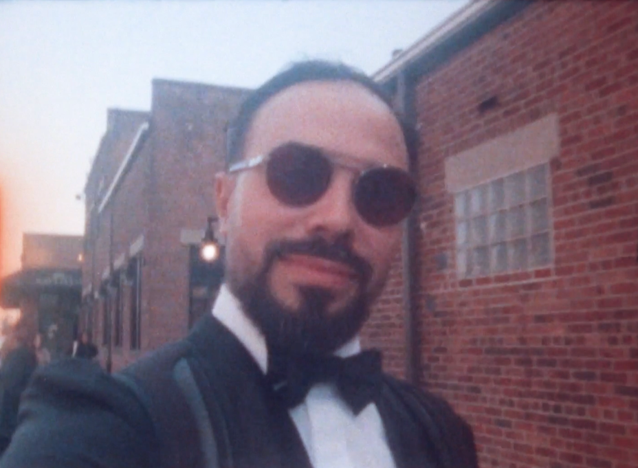 A man with a beard and sunglasses taking a selfie outdoors at sunset, dressed in a tuxedo with a bow tie, standing against a brick wall.