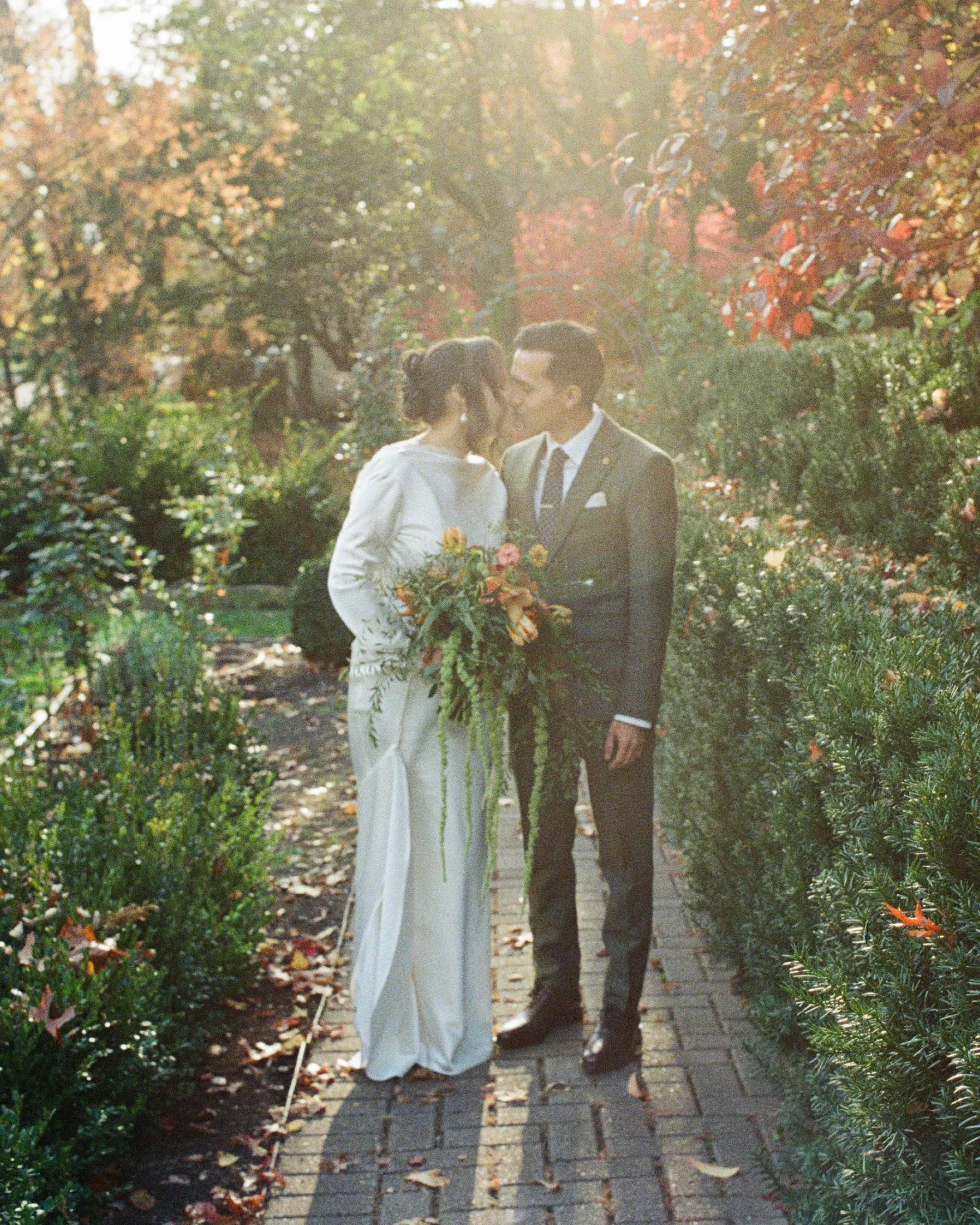 A couple dressed in wedding attire walking on a garden path surrounded by trees with autumn foliage, with sunlight filtering through the leaves.