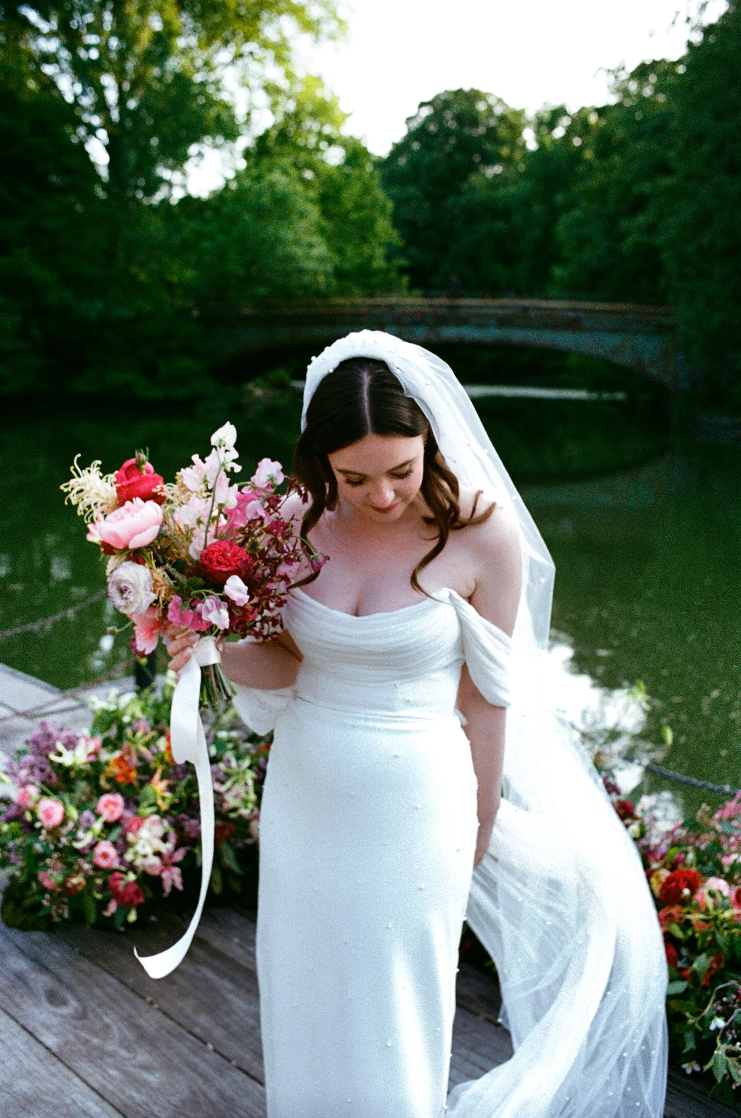 A bride in a white wedding dress holding a bouquet of pink and red flowers, standing on a wooden dock by a body of water with trees and a bridge in the background.