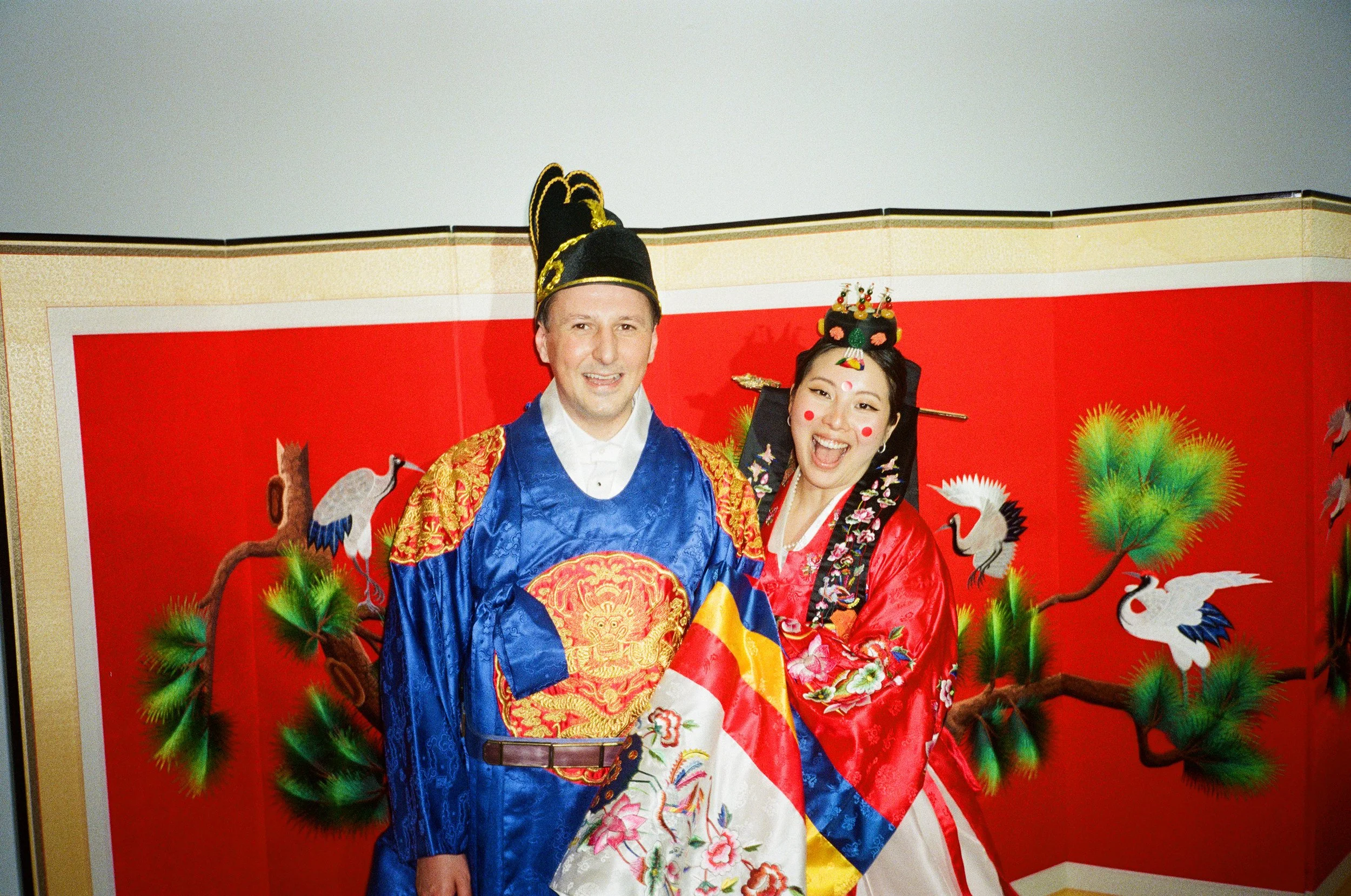 Two people dressed in traditional Korean costumes, standing in front of a red backdrop decorated with cranes and pine trees.