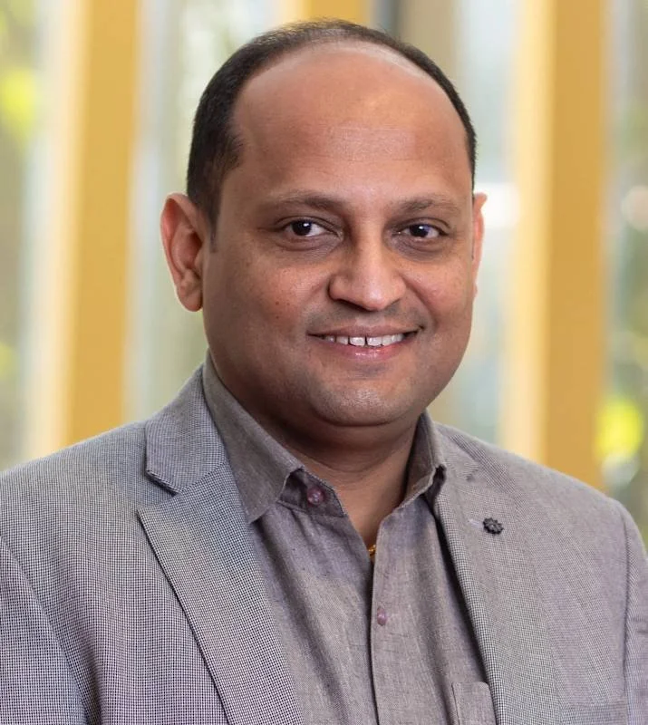 A man with a shaved head wearing a light grey blazer and a button-up shirt, smiling, standing in front of large windows with a blurry outdoor background.