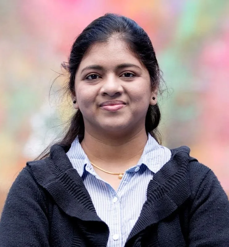A woman standing outdoors with her arms crossed, wearing a black sweater over a blue and white striped shirt, with a blurred colorful background.