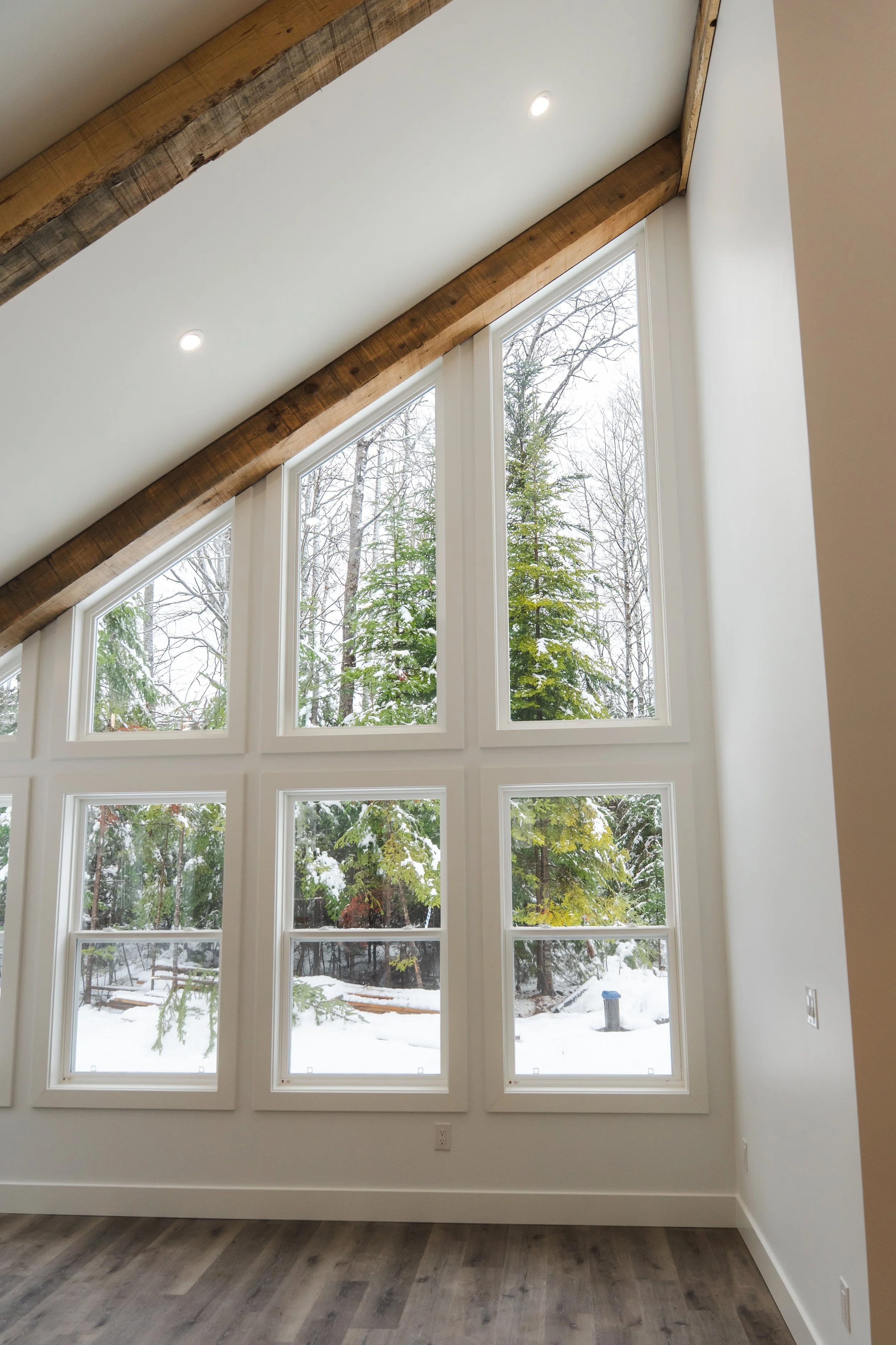 A room with large, multi-pane windows showing a snowy outdoor scene with trees, a bench, and a trash can. The ceiling has recessed lighting and a wooden beam.