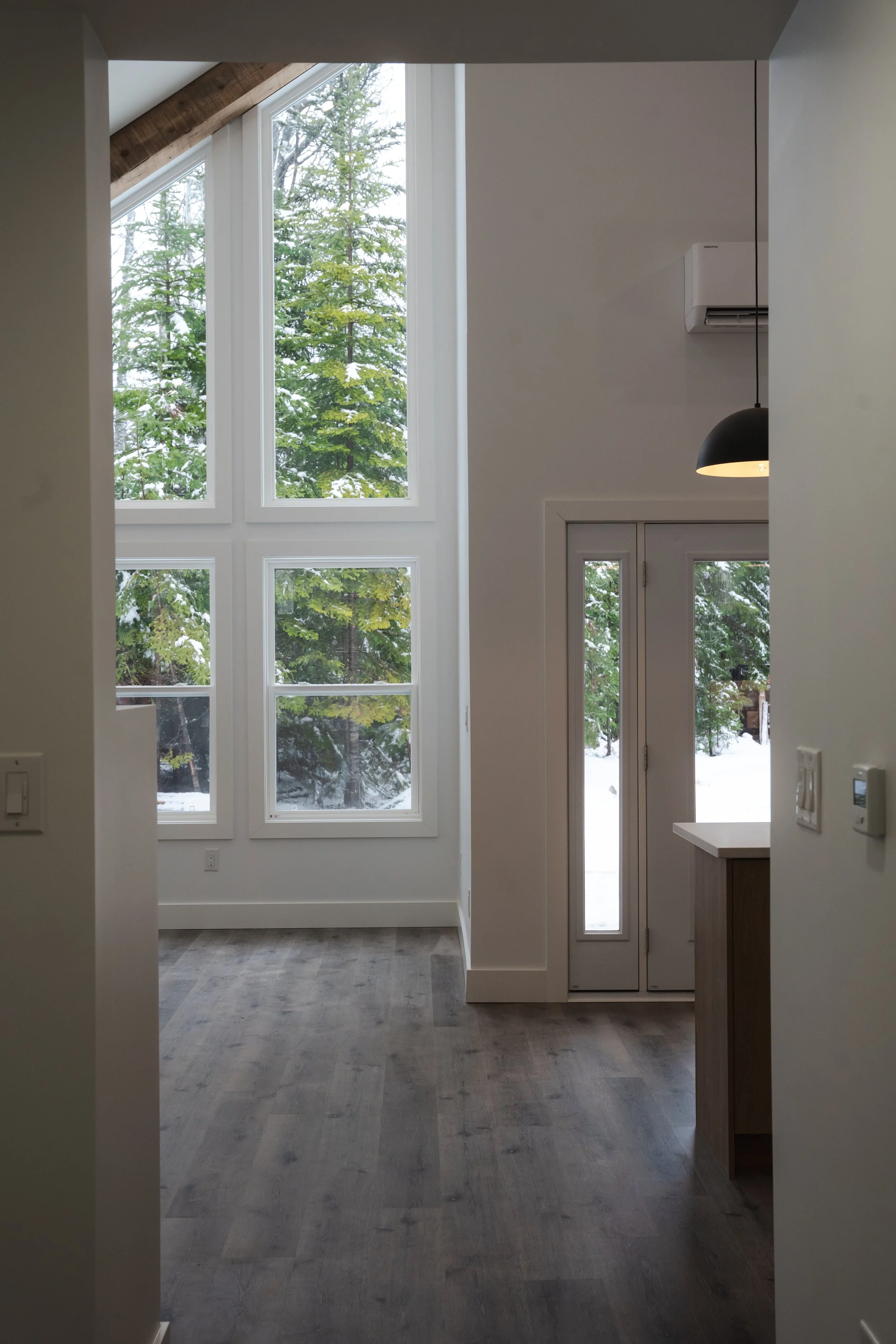 Interior view of a modern room with large windows showing snow-covered trees outside, a door leading outside, and hardwood flooring.