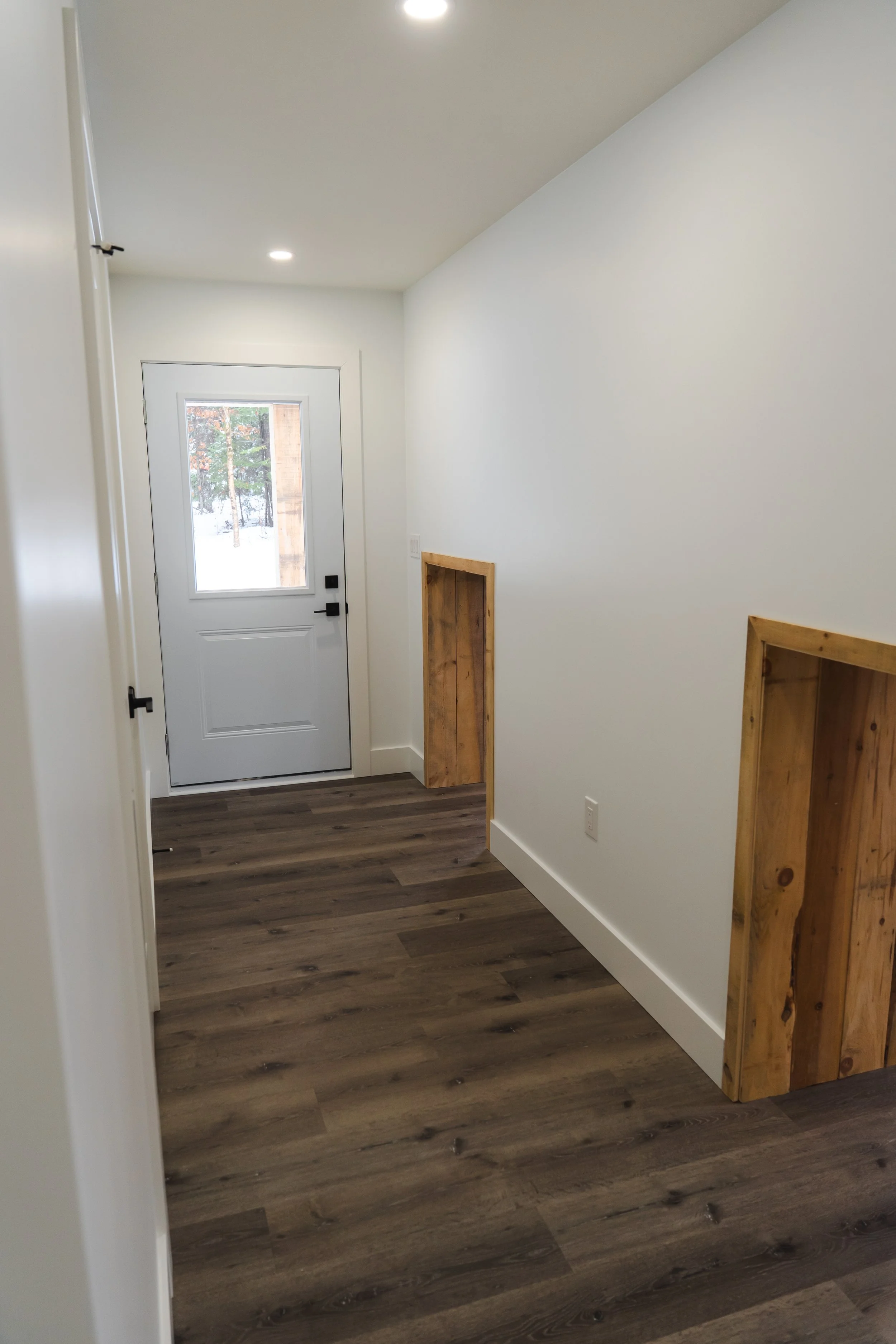 Entryway of a house with white walls, dark wood flooring, a white door with a window, and two built-in wall niches framed with wood.