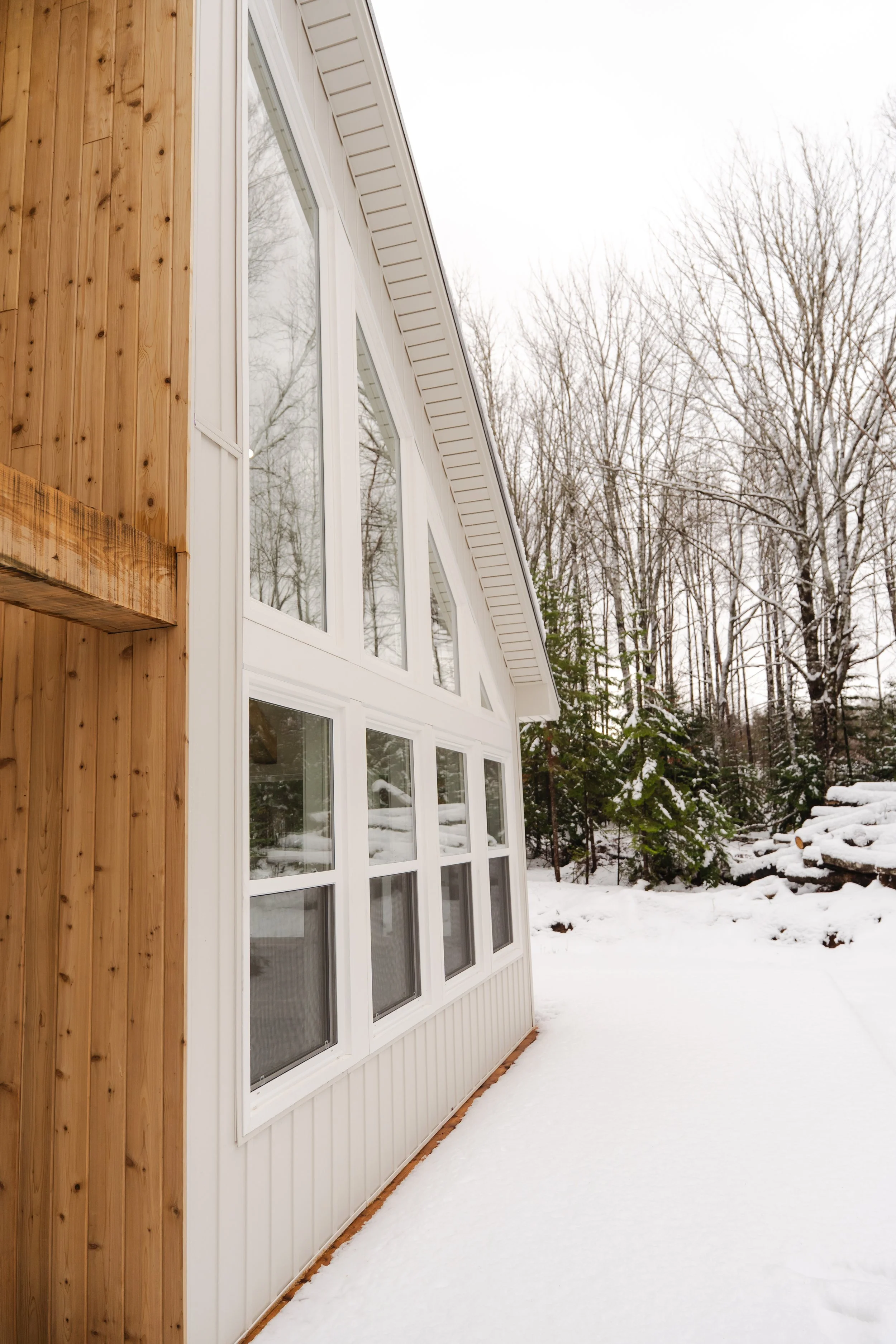Side view of a house with large windows, wood siding on one part and white siding on the other, snow-covered ground, and snow-laden trees in the background.