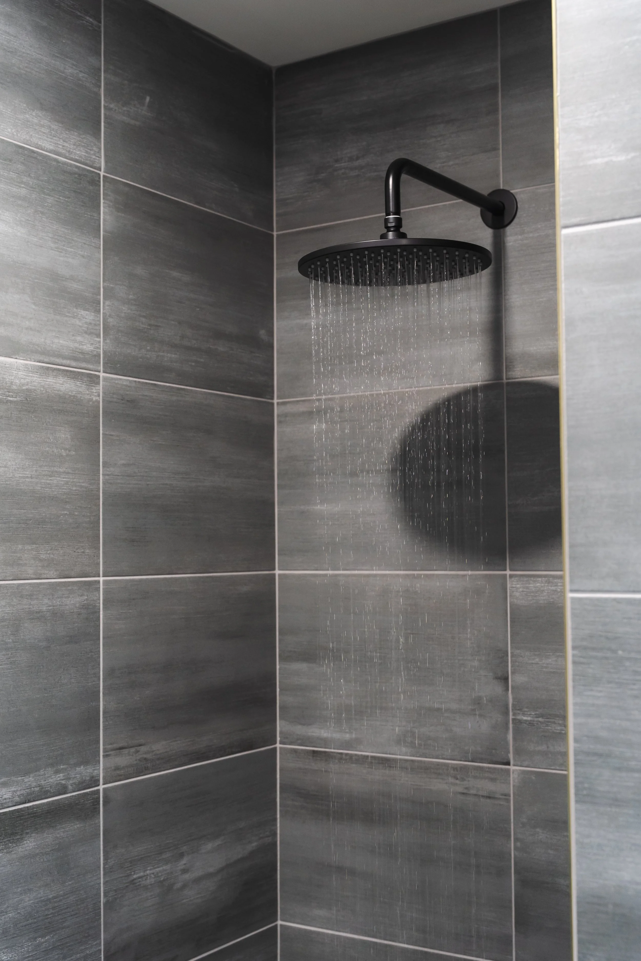 A black showerhead mounted on a wall, with water flowing down, in a bathroom with gray tiled walls.
