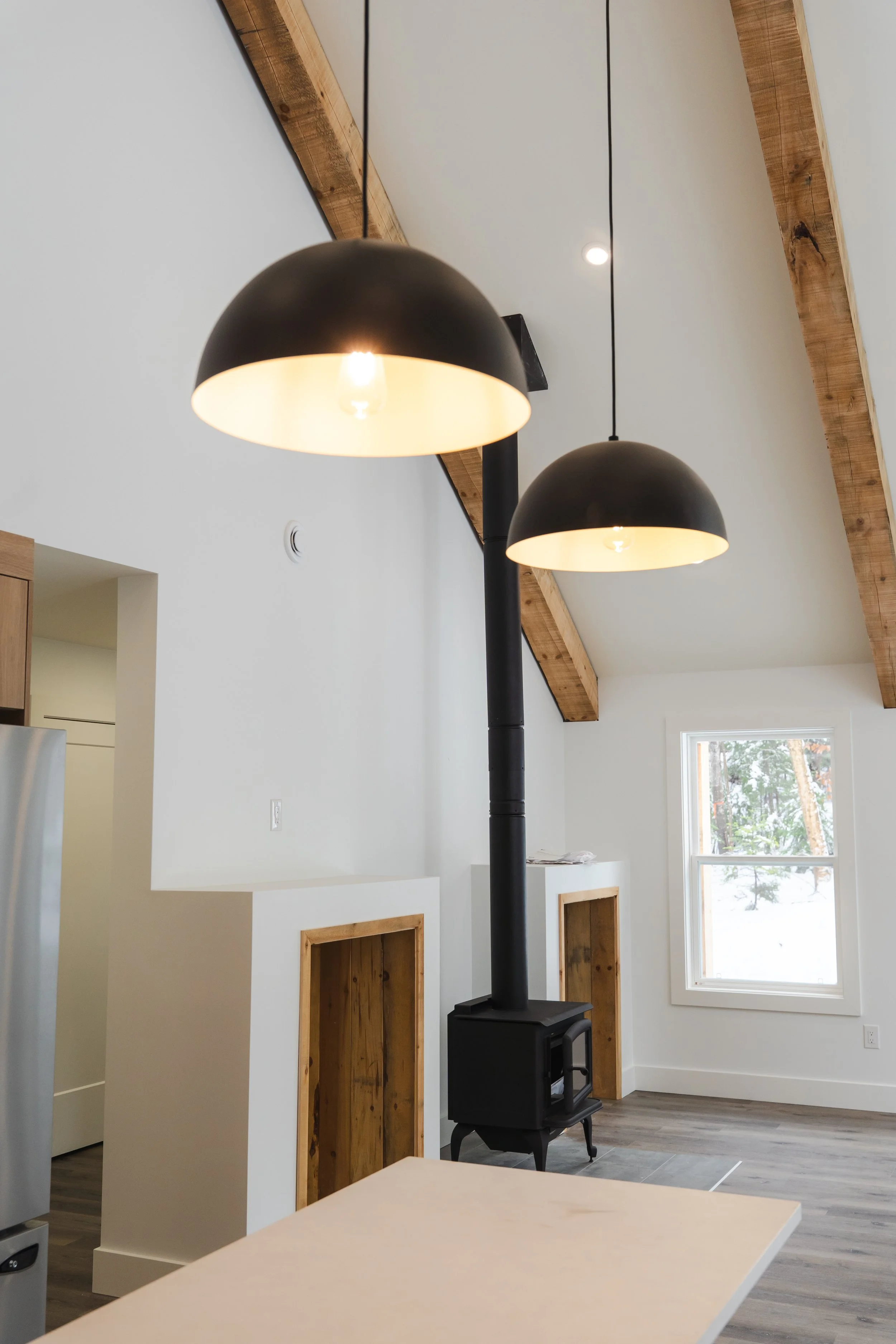 Interior of a modern home with two black dome pendant lights hanging from a ceiling with wooden beams. A wood stove is against a white wall, and there is a window showing trees outside.