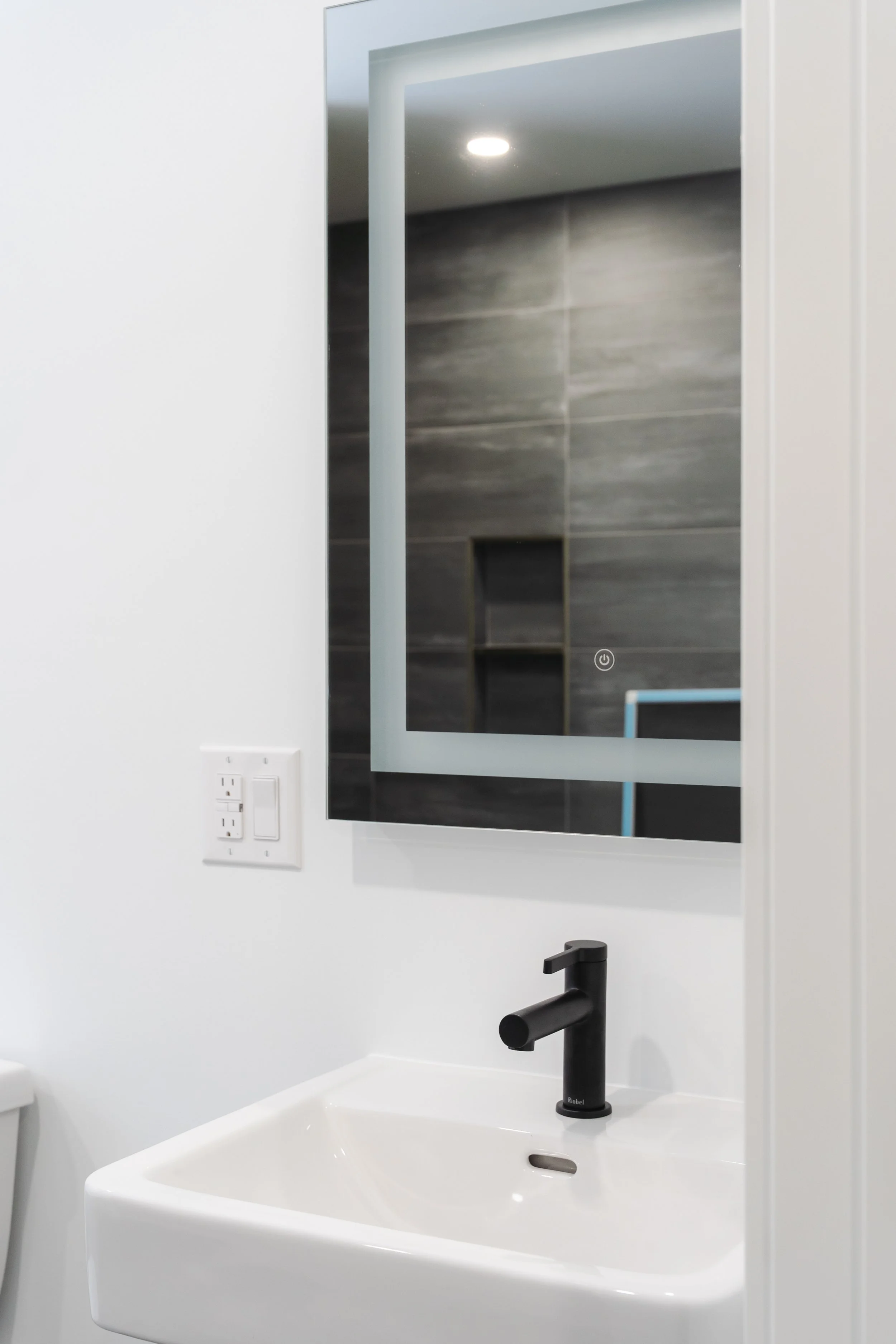 A bathroom with a white vanity sink, black faucet, large mirror, and gray tiled shower visible in the reflection.
