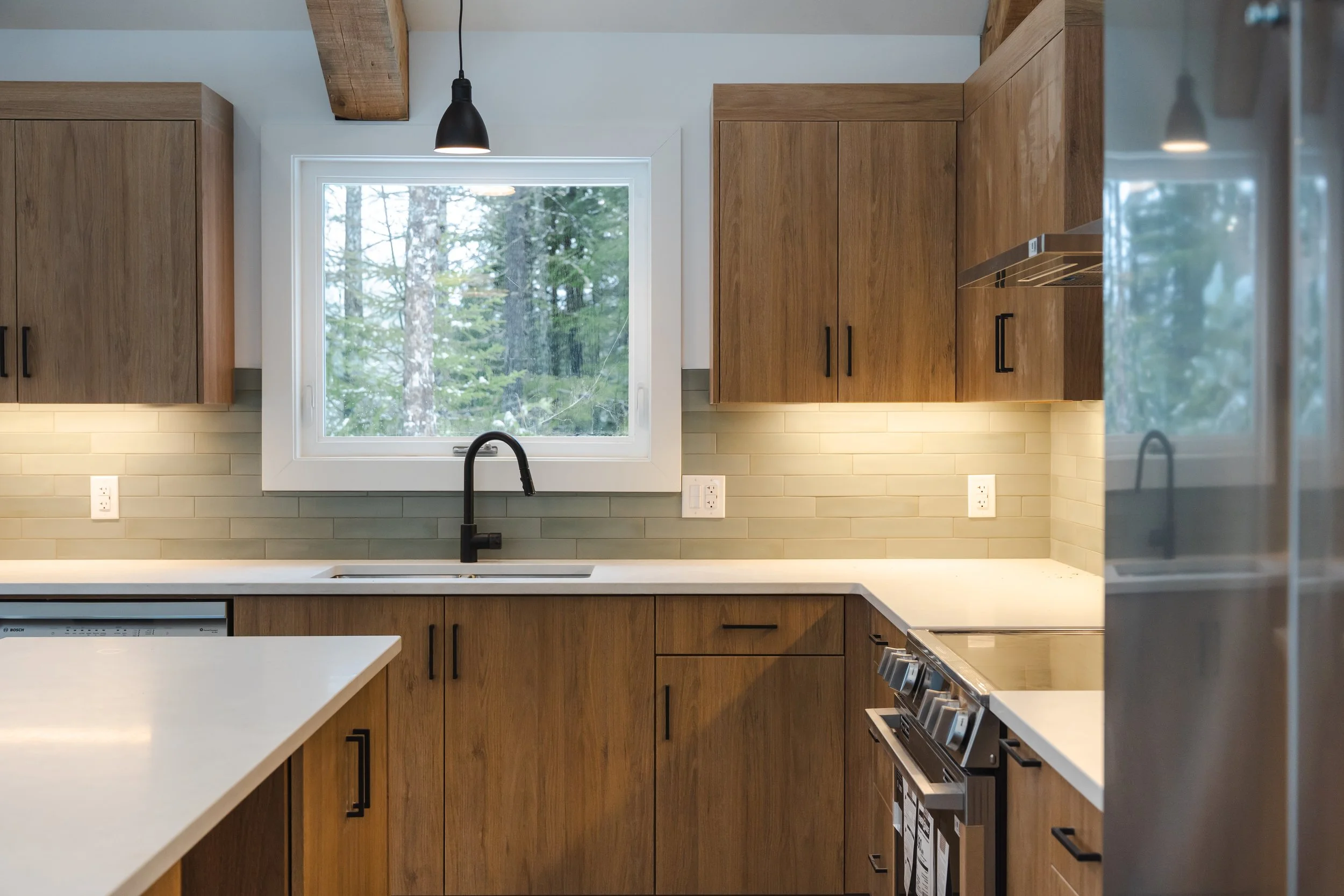 Modern kitchen with wooden cabinets, white countertops, a large window showing an outdoor view of trees, black faucet, and stainless steel appliances.