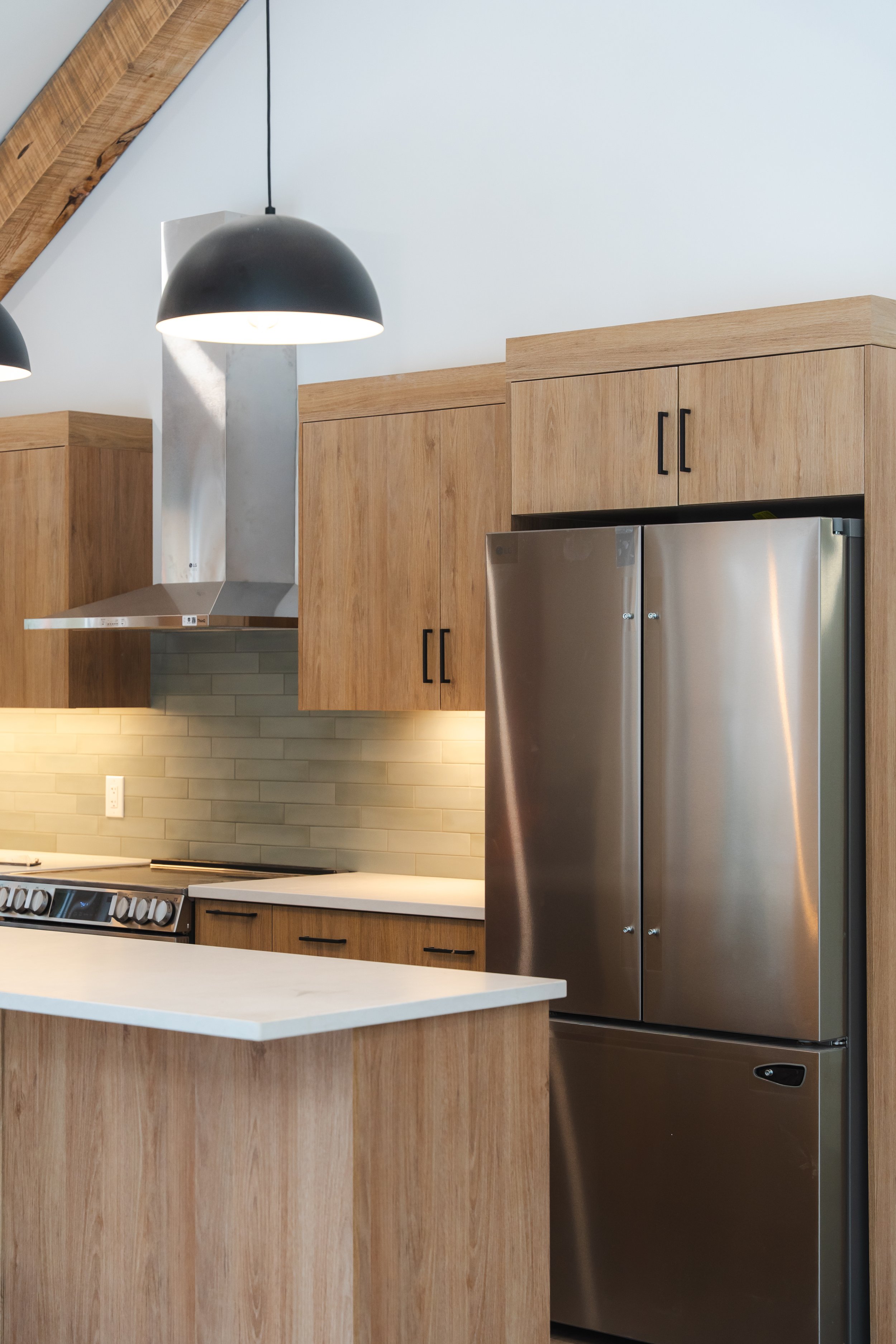 Modern kitchen with wooden cabinets, stainless steel refrigerator, range stove, subway tile backsplash, and black pendant lights.