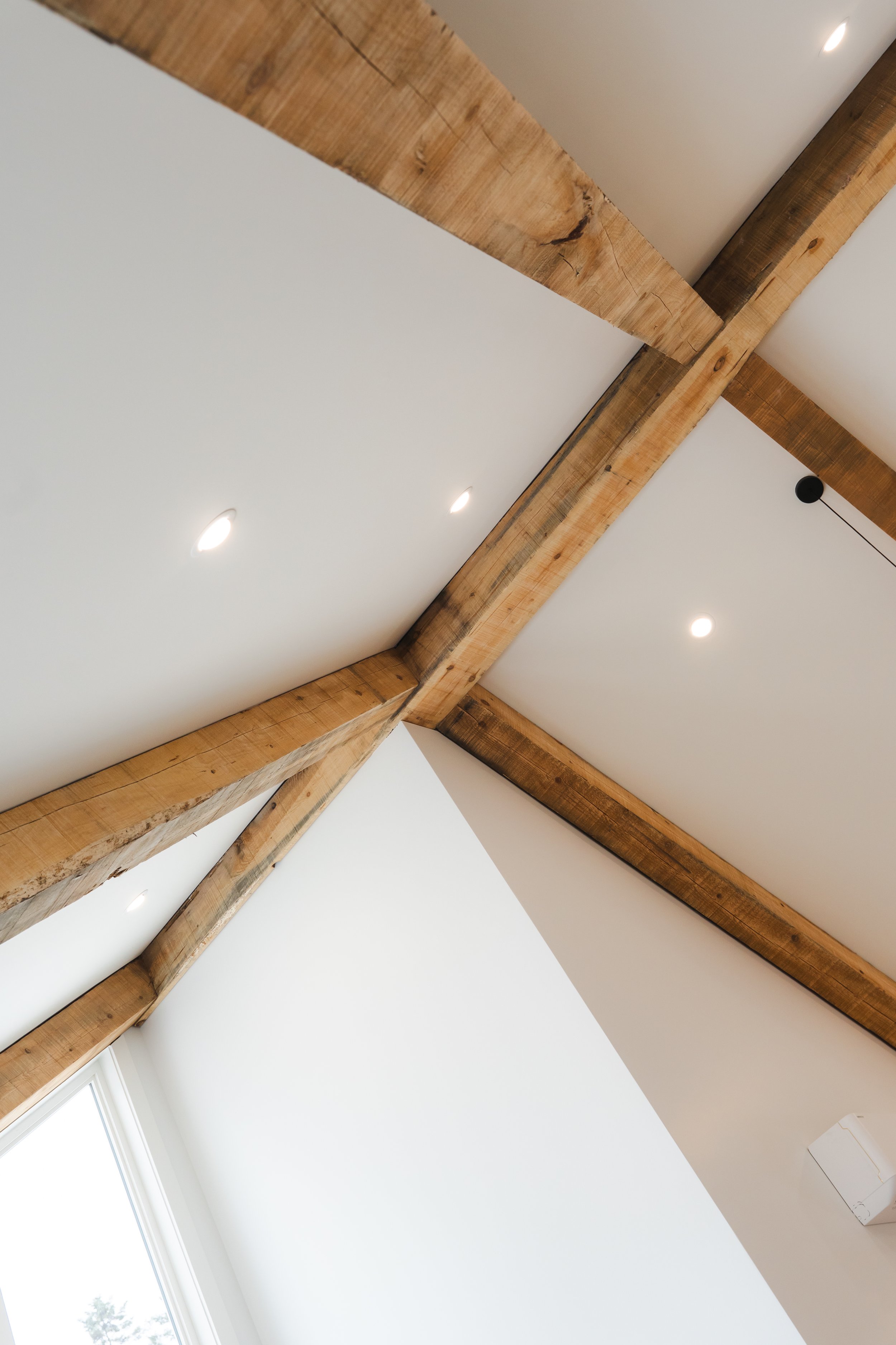 Interior ceiling with exposed wooden beams, recessed lighting, and a portion of a window.