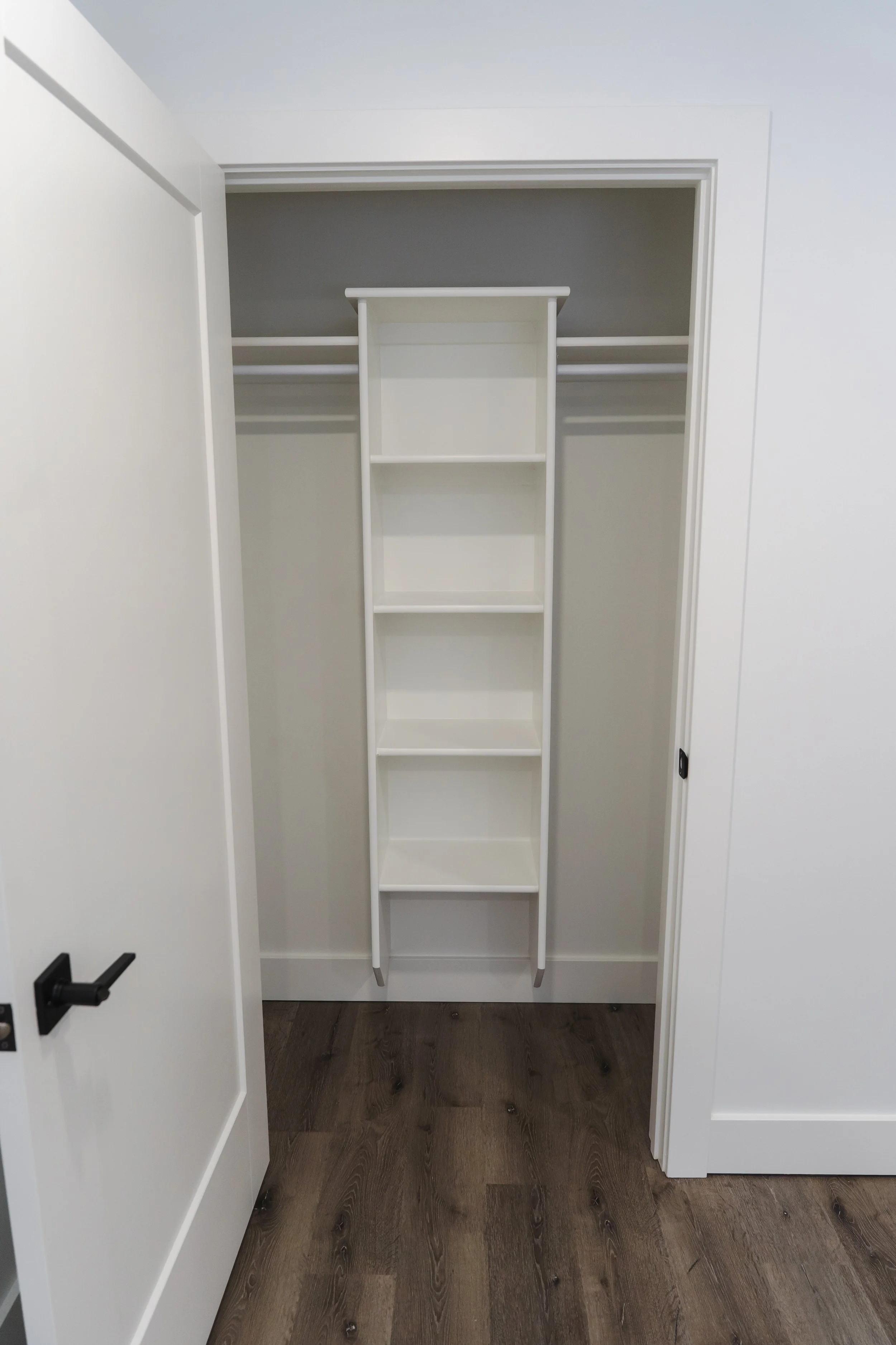 Empty closet with a white ladder shelf, hanging rods on both sides, white walls, and wood flooring.