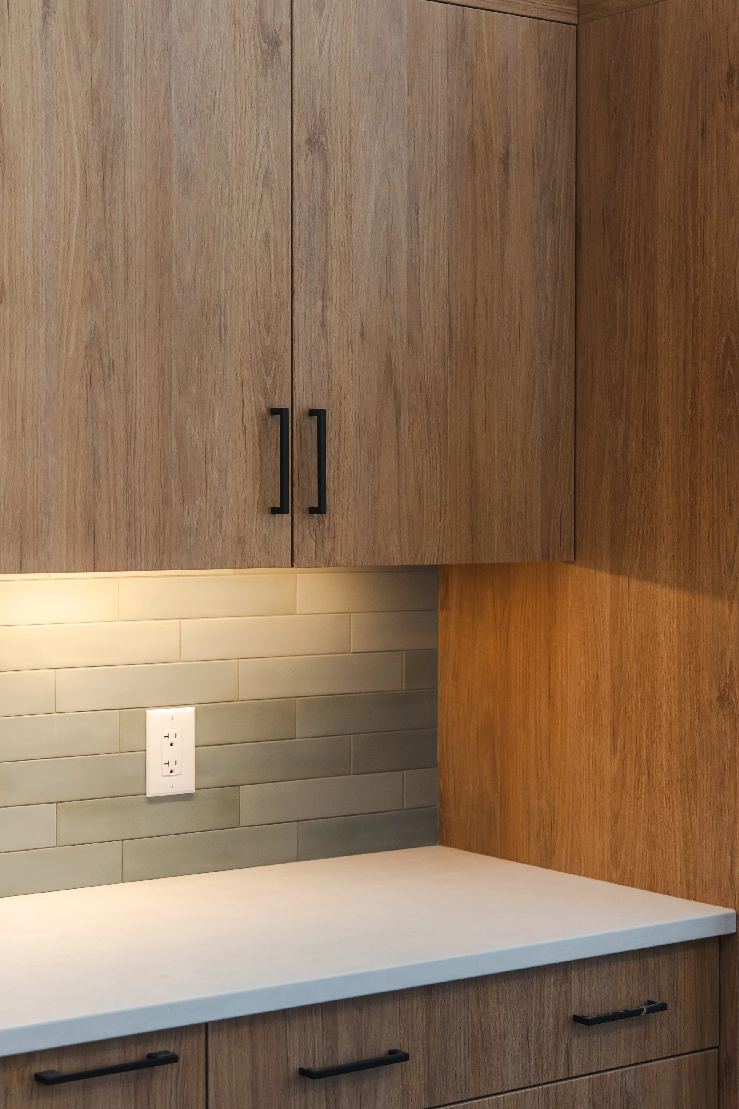 Kitchen counter with wooden cabinets, a beige countertop, gray tiled backsplash, and a wall outlet.