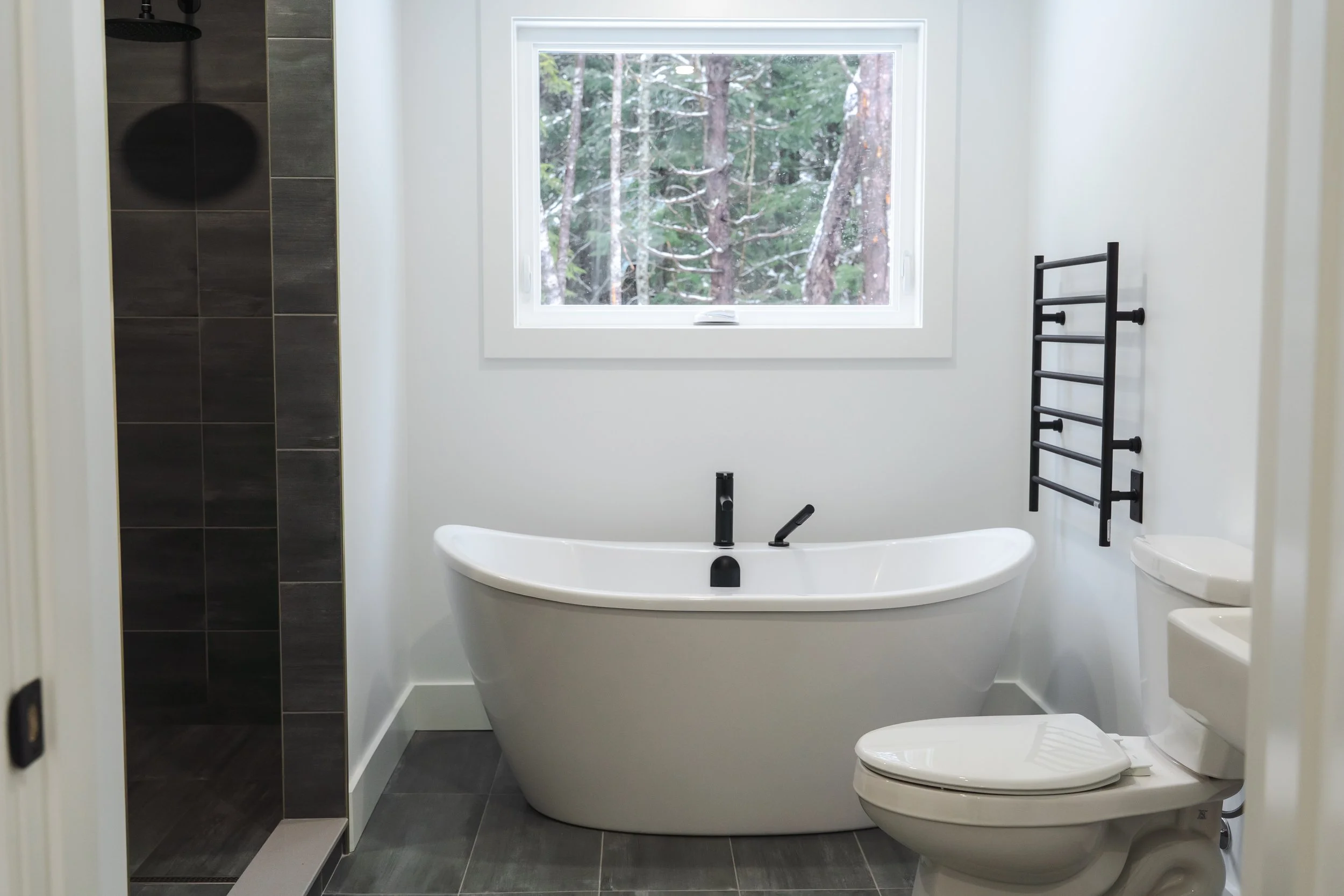 Modern bathroom with a walk-in shower with black tiles, a white freestanding bathtub, a toilet, a black towel rack, and a large window showing a view of trees.