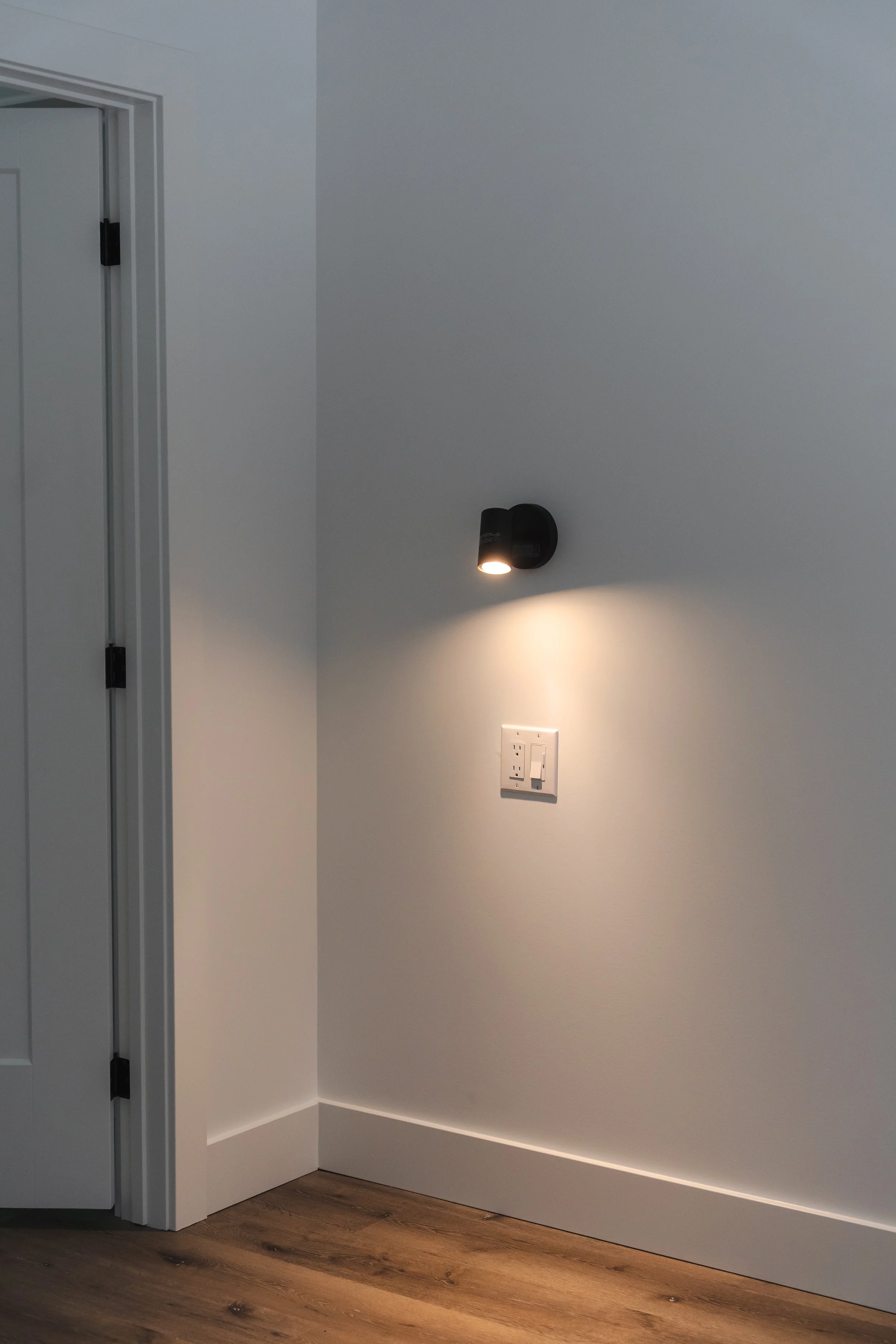 A wall with a mounted black light fixture turned on, beside a white electrical outlet and switch plate, with a wooden floor at the bottom and a partially open door to the left.