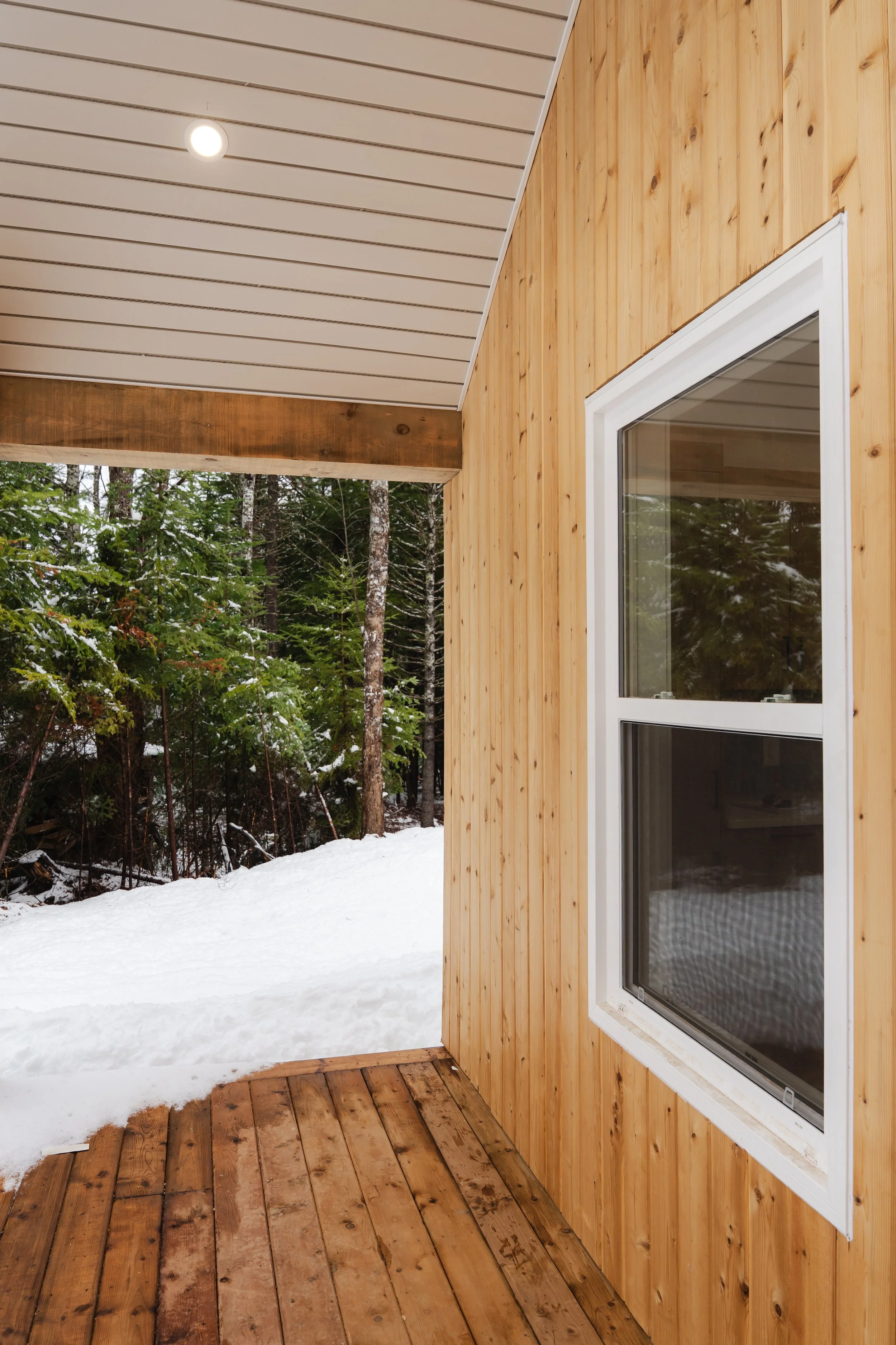 View of a wooden porch next to a house with a window, overlooking a snowy landscape with trees.