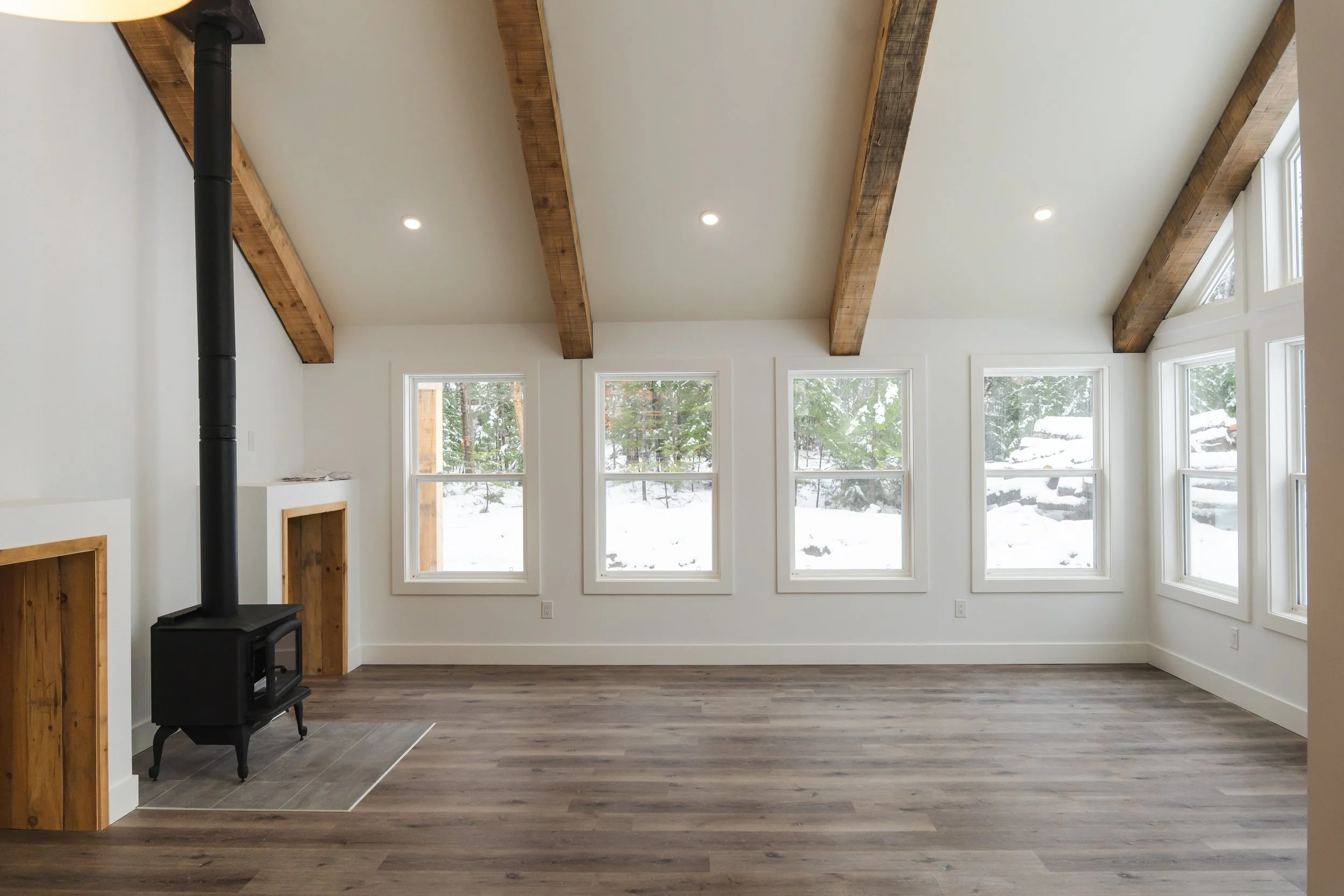 Empty living room with white walls, wooden beams on the ceiling, a wood stove in the corner, large windows showing snowy outdoors, dark wood flooring.
