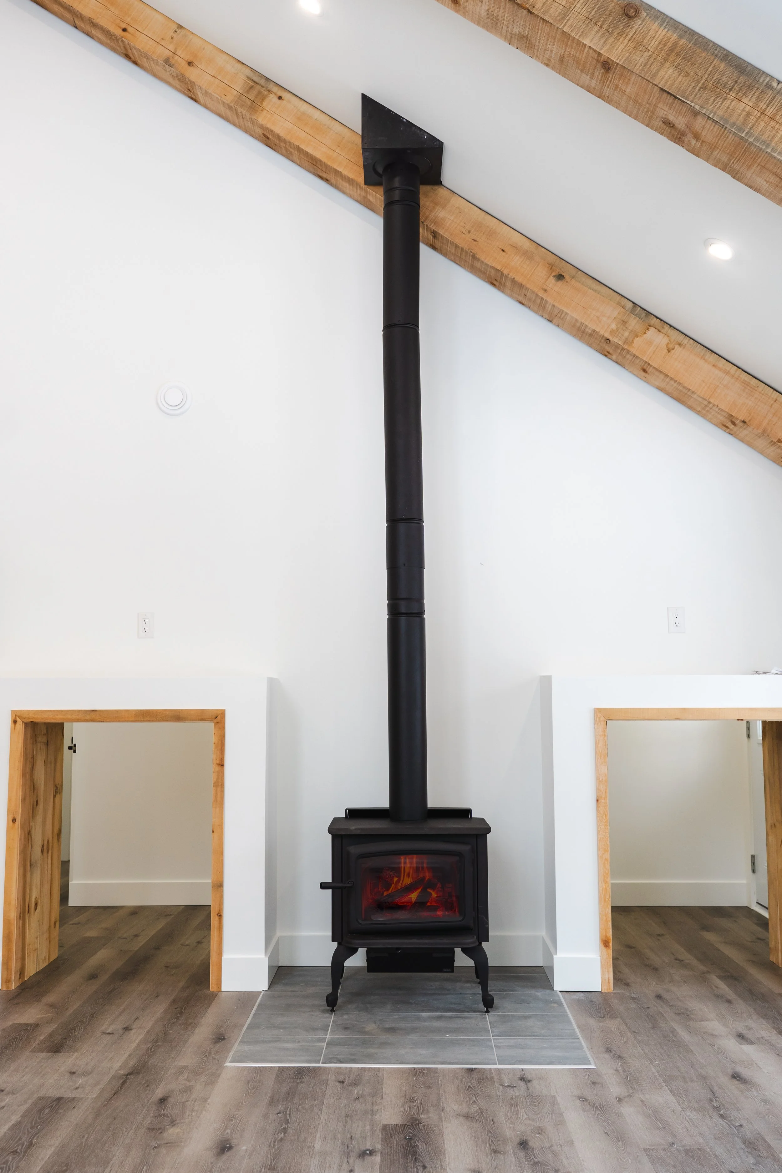 A black wood stove with a chimney pipe in a white room with wooden beams and trim, and hardwood floors.