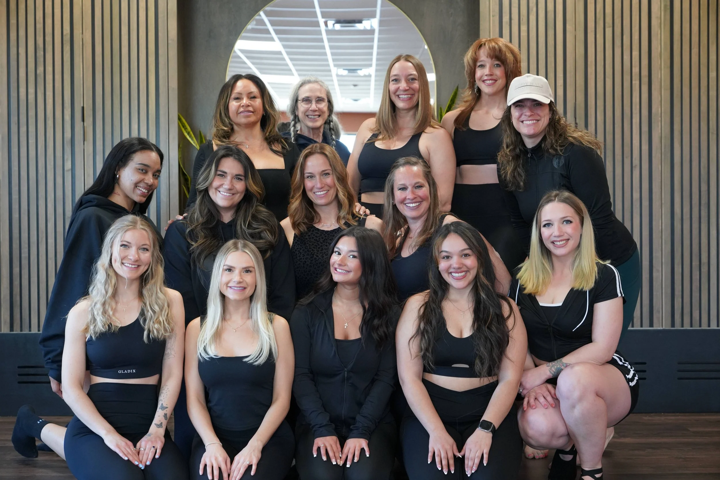 A diverse group of women posing together indoors, smiling, with wooden paneling and plants in the background.