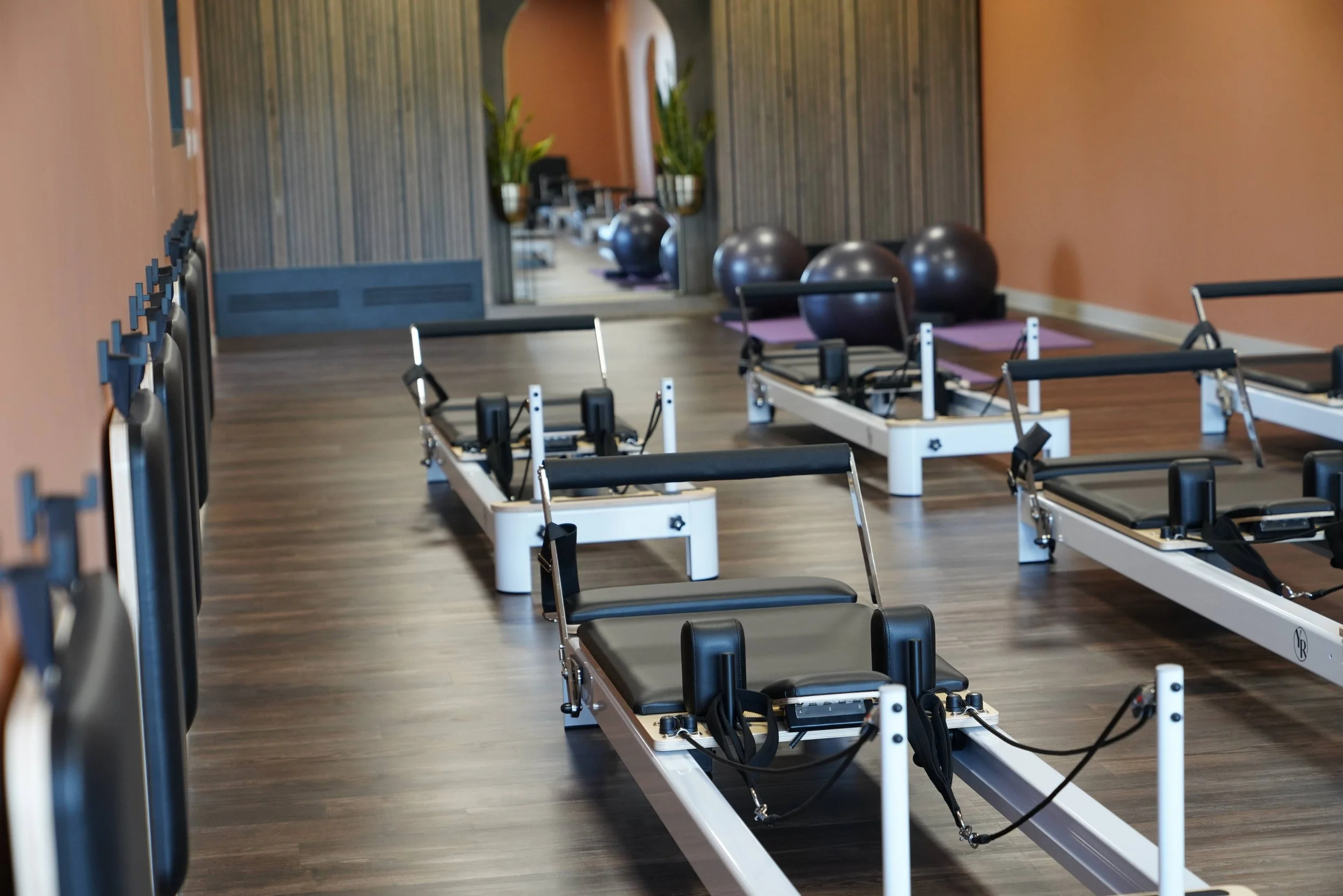 Fitness studio with Pilates reformer machines, exercise balls, and stability chairs, with a mirrored wall and plants in the background.