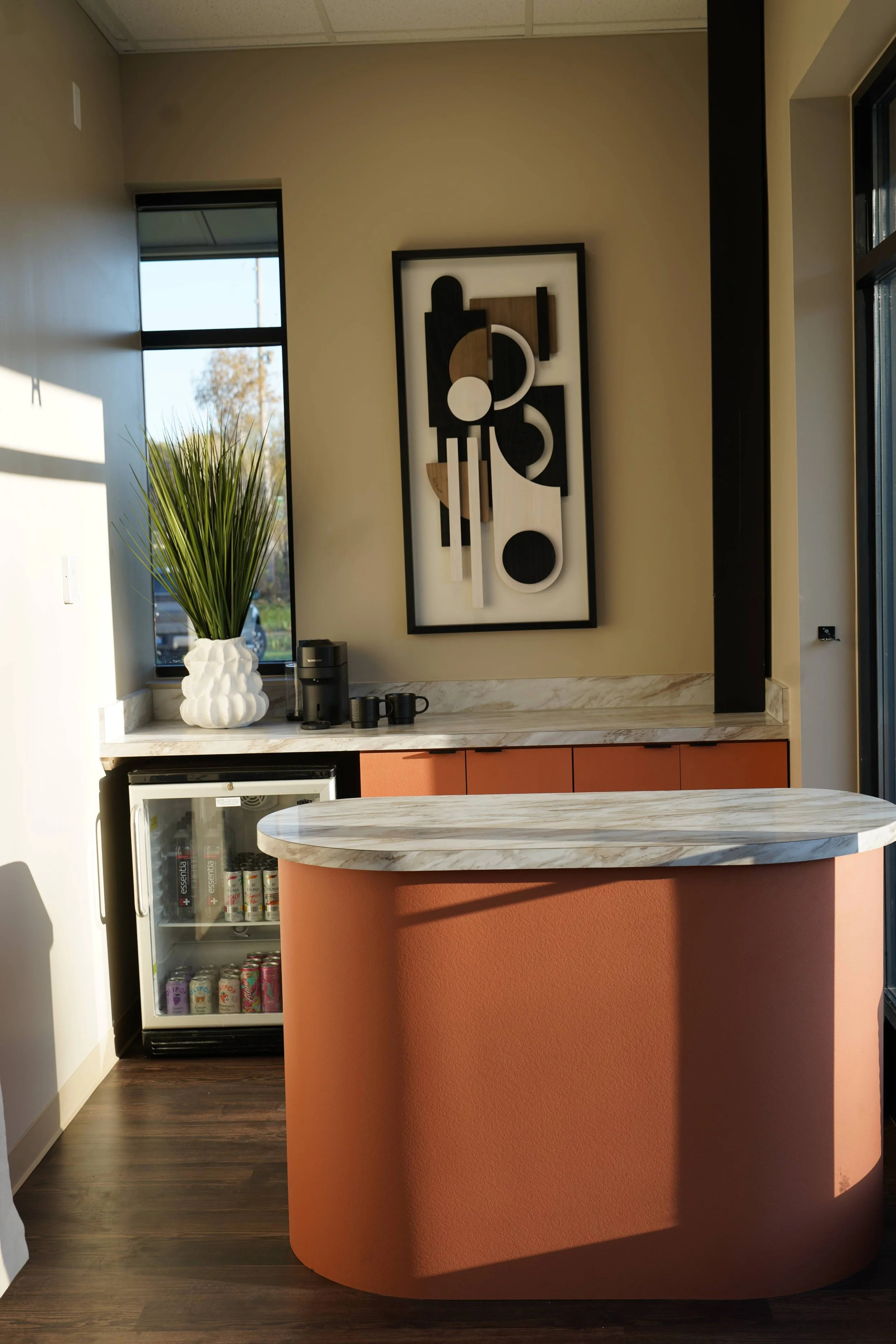 A BHLC countertop with a decorative vase with green plants, a coffee machine, and cups. Behind it is a framed abstract black, white, and brown artwork on a beige wall.