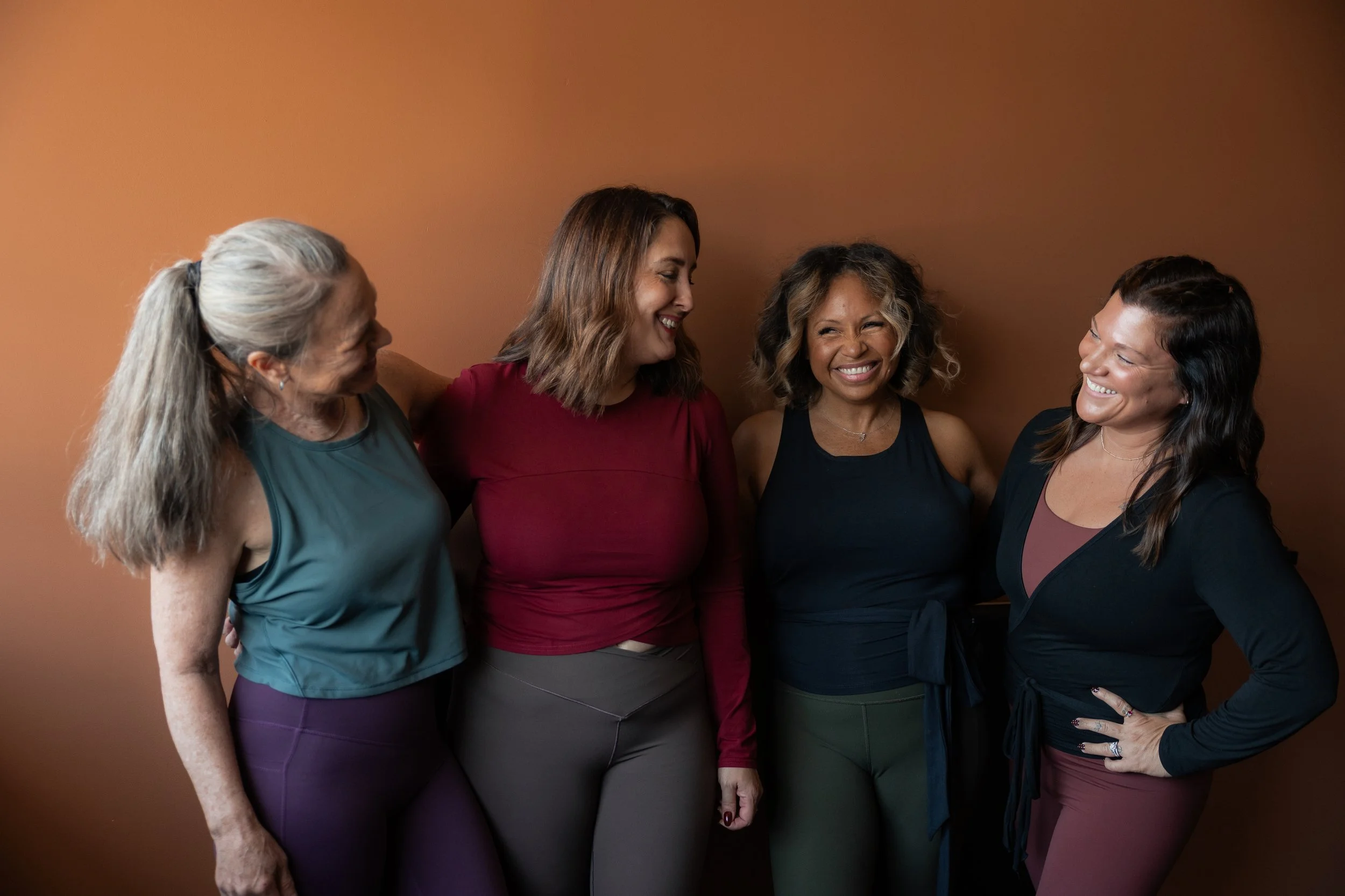 Four women are standing together, smiling and looking at each other in front of a plain brown background.