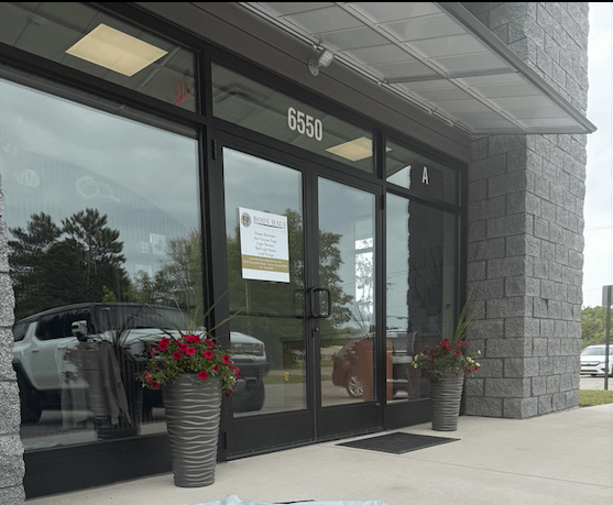 The entrance of a commercial building with glass doors and the number '6550' above it, flanked by two large potted plants with flowers.