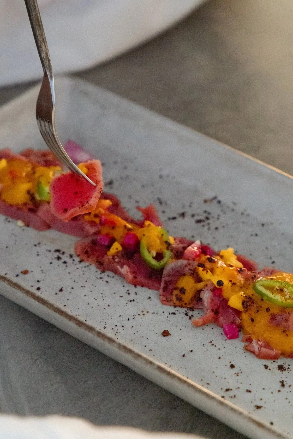 Close-up of a person using a fork to pick up a piece of sashimi with colorful toppings on a rectangular plate.