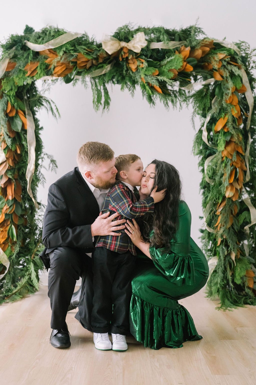 A man and woman in formal attire kneeling with a child in front of them, under a decorative arch of green foliage and ribbons, indoors on a light wood floor.