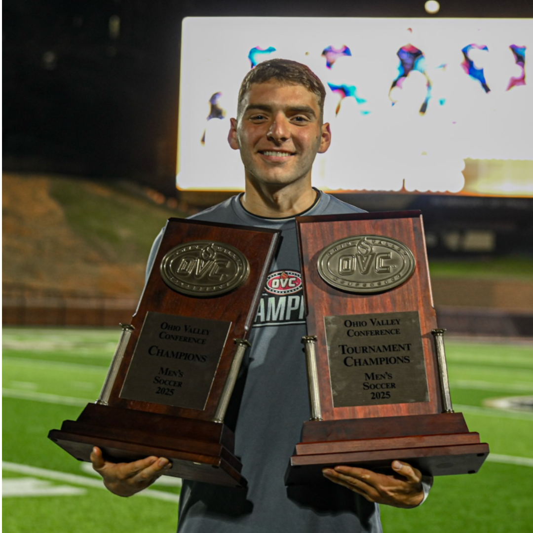 A young man smiling on a soccer field, holding two Ohio Valley Conference tournament championship plaques for men's soccer, 2025.