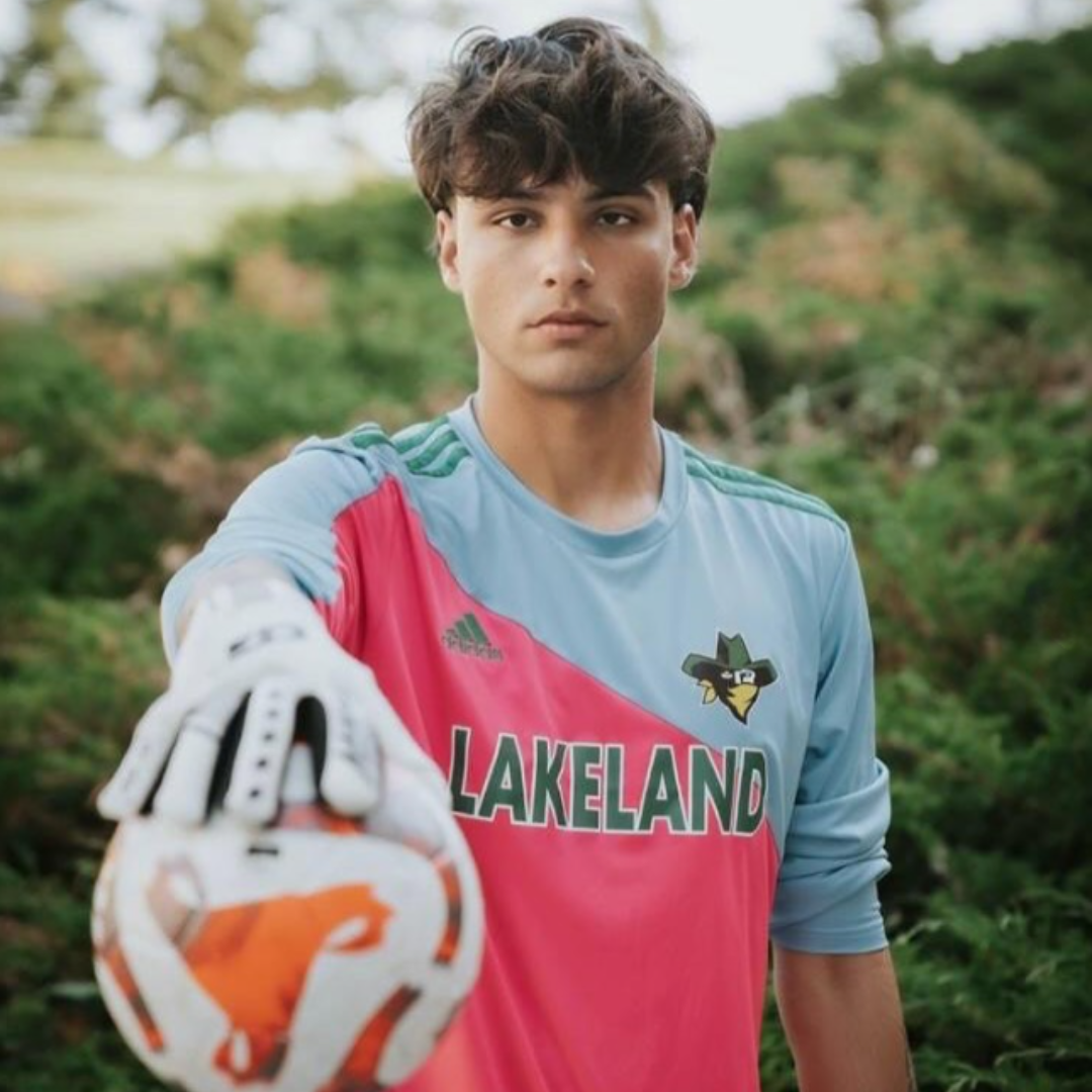 Young male soccer goalkeeper wearing a pink and blue jersey with green accents, extending his gloved hand towards the camera with a soccer ball for a close-up shot, outdoors in a lush green setting.