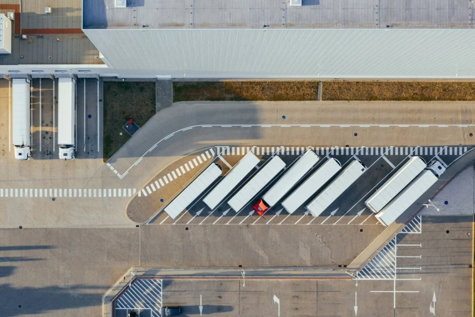 Aerial view of a logistics center with a row of docked white semi-trailer trucks, the loading bays under a large metal roof, with an adjacent parking lot and road, creating an organized industrial pattern.