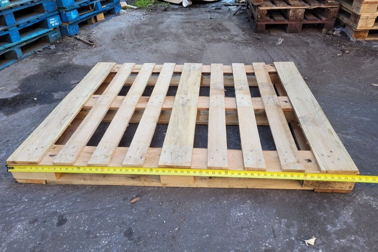 A wooden pallet with a tape measure stretched across its width, situated outdoors on a concrete surface, with other pallets stacked in the background.