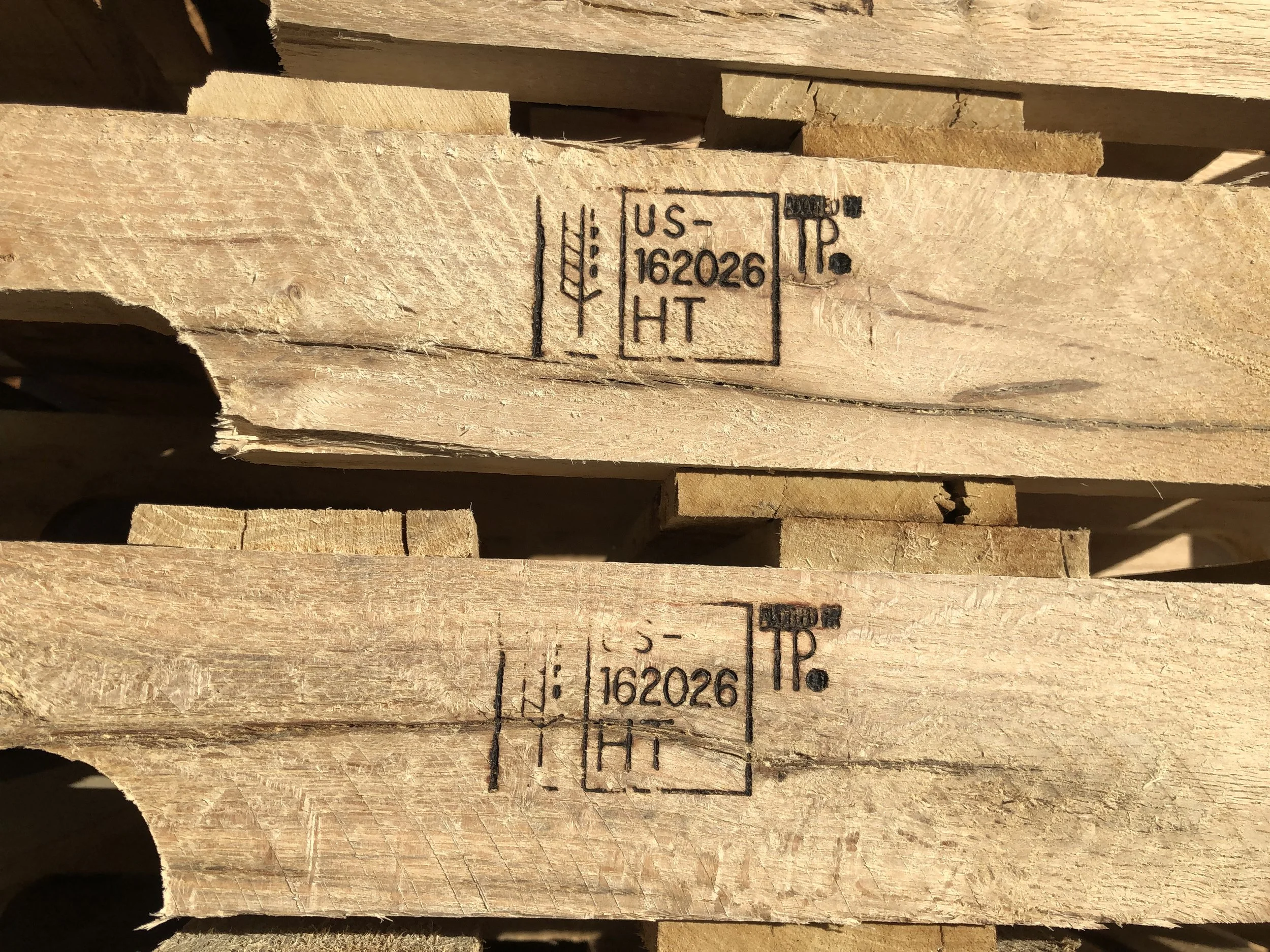 A stack of natural wooden pallets with visible grain, featuring a heat treatment stamp on the side blocks marked with "HT" inside a rectangular outline, and additional markings indicating certification for international shipping