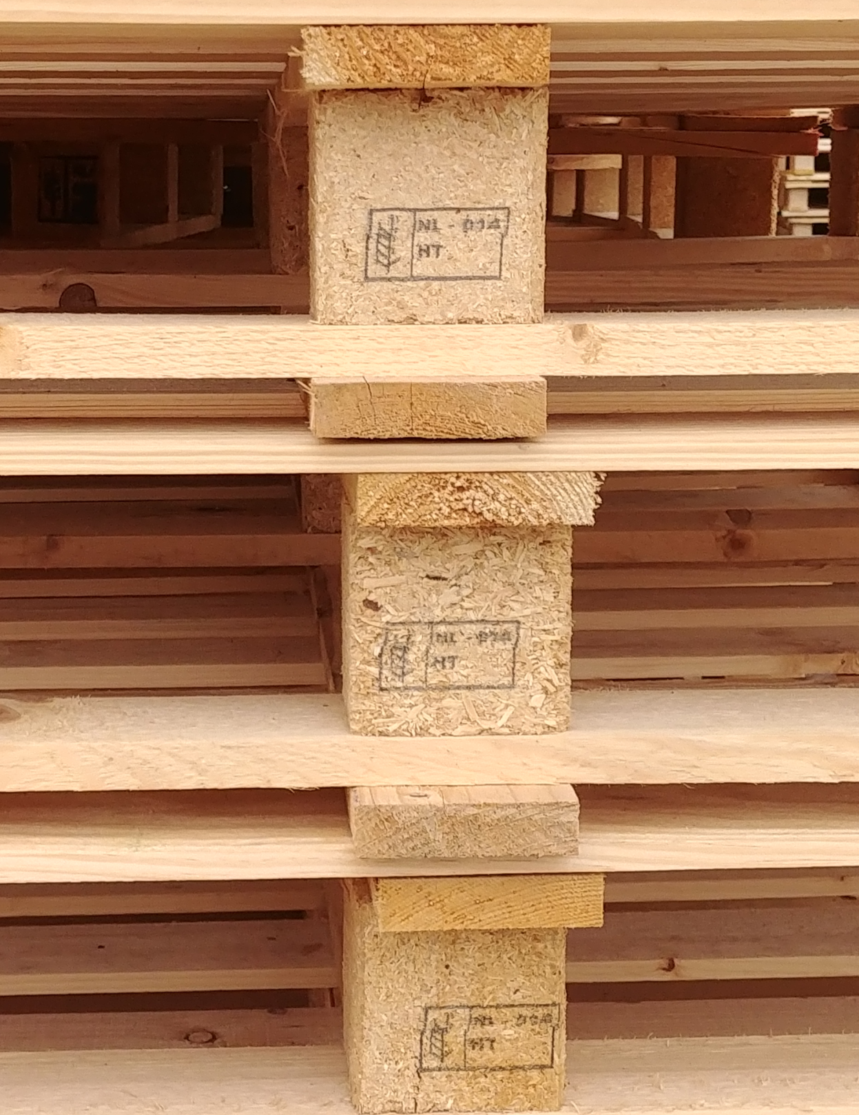 A stack of natural wooden pallets with visible grain, featuring a heat treatment stamp on the side blocks marked with "HT" inside a rectangular outline, and additional markings indicating certification for international shipping