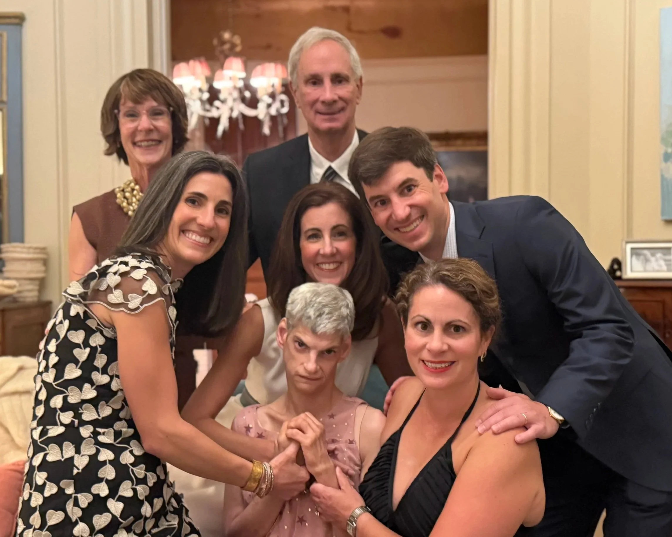 Group of eight people, six women and two men, gathered in a cozy, elegant room, posing for a photograph, smiling, with warm lighting and decorative background.