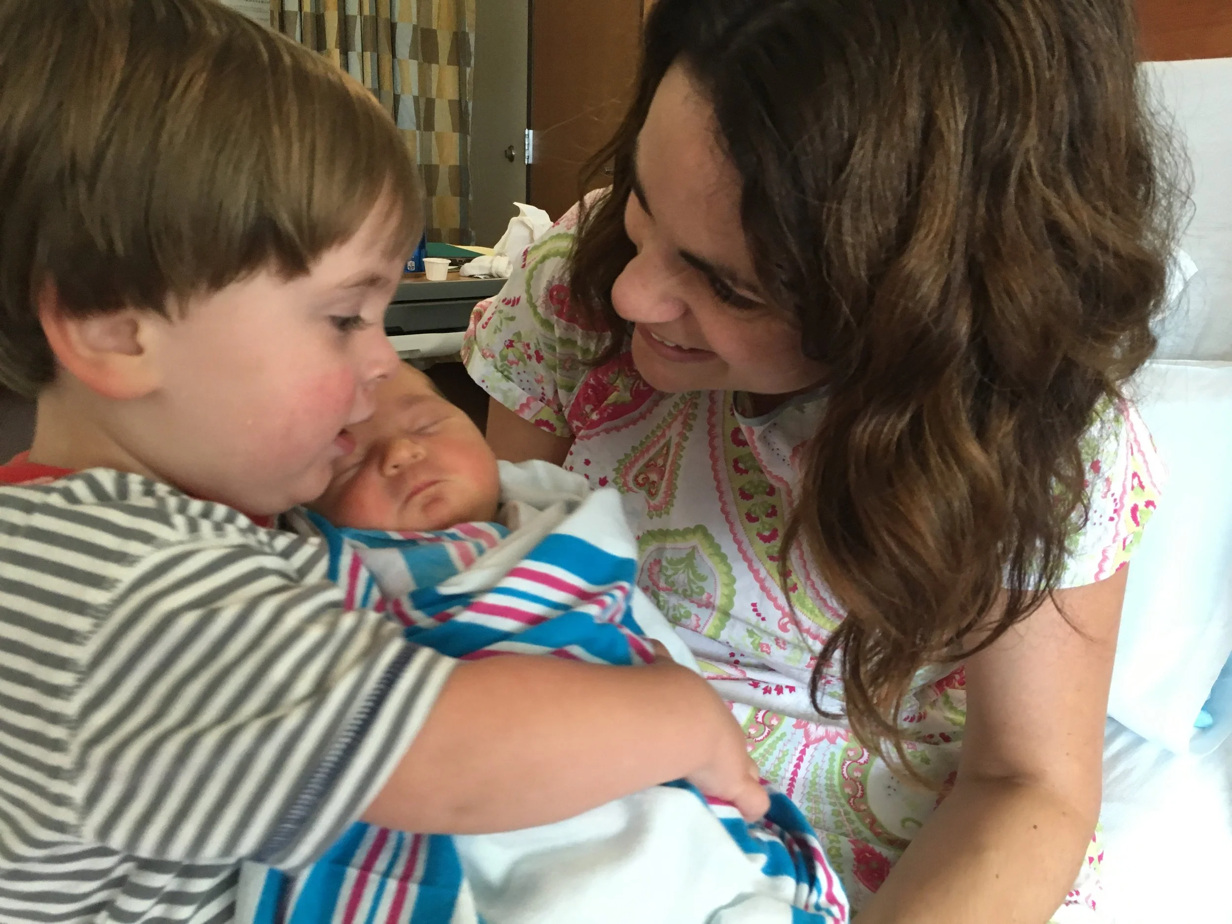 A young boy holding a newborn baby in a hospital room, smiling as the woman looks affectionately at them.