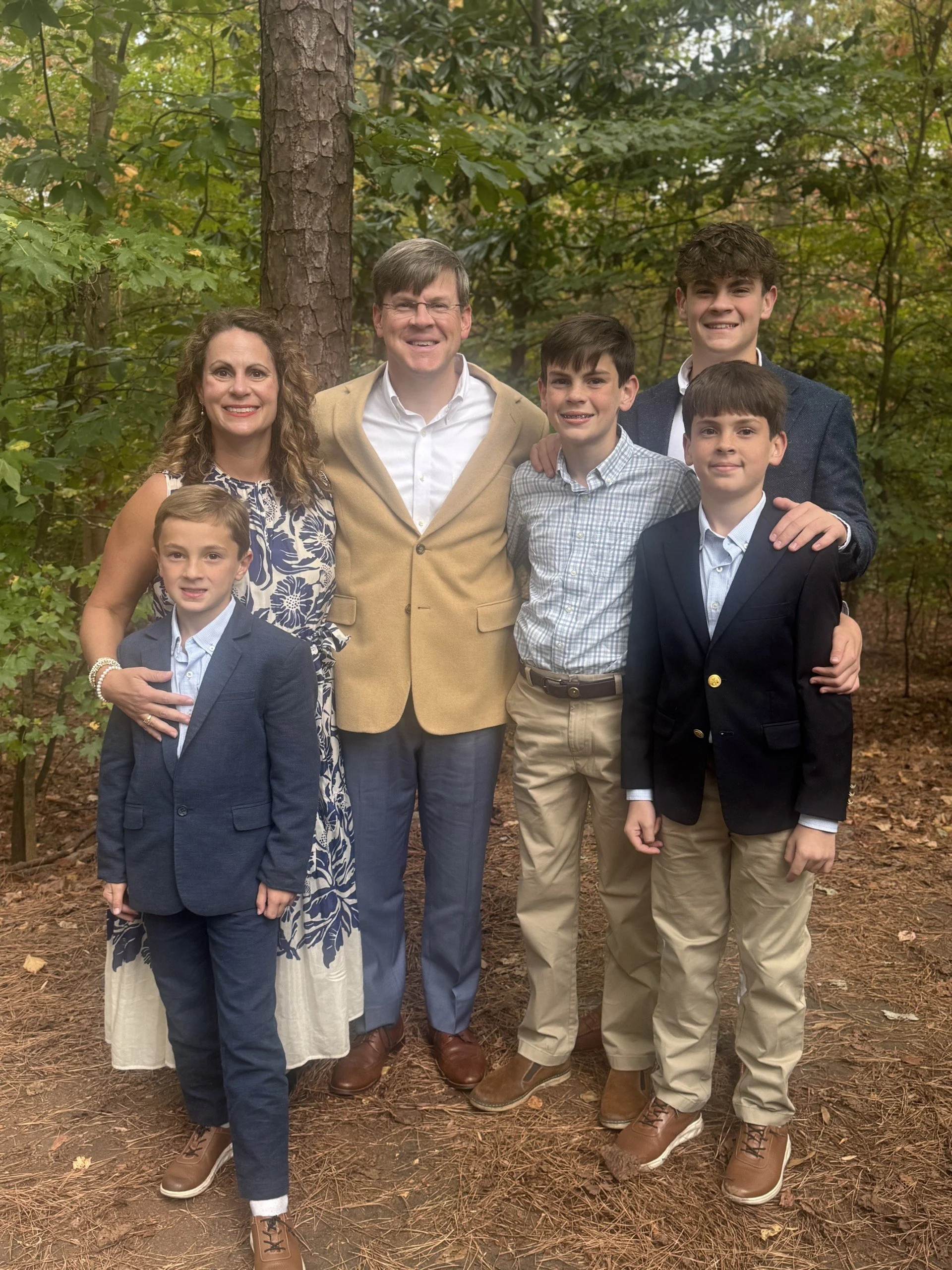 A family of seven posing outdoors in a wooded area, dressed in semi-formal attire, smiling at the camera.