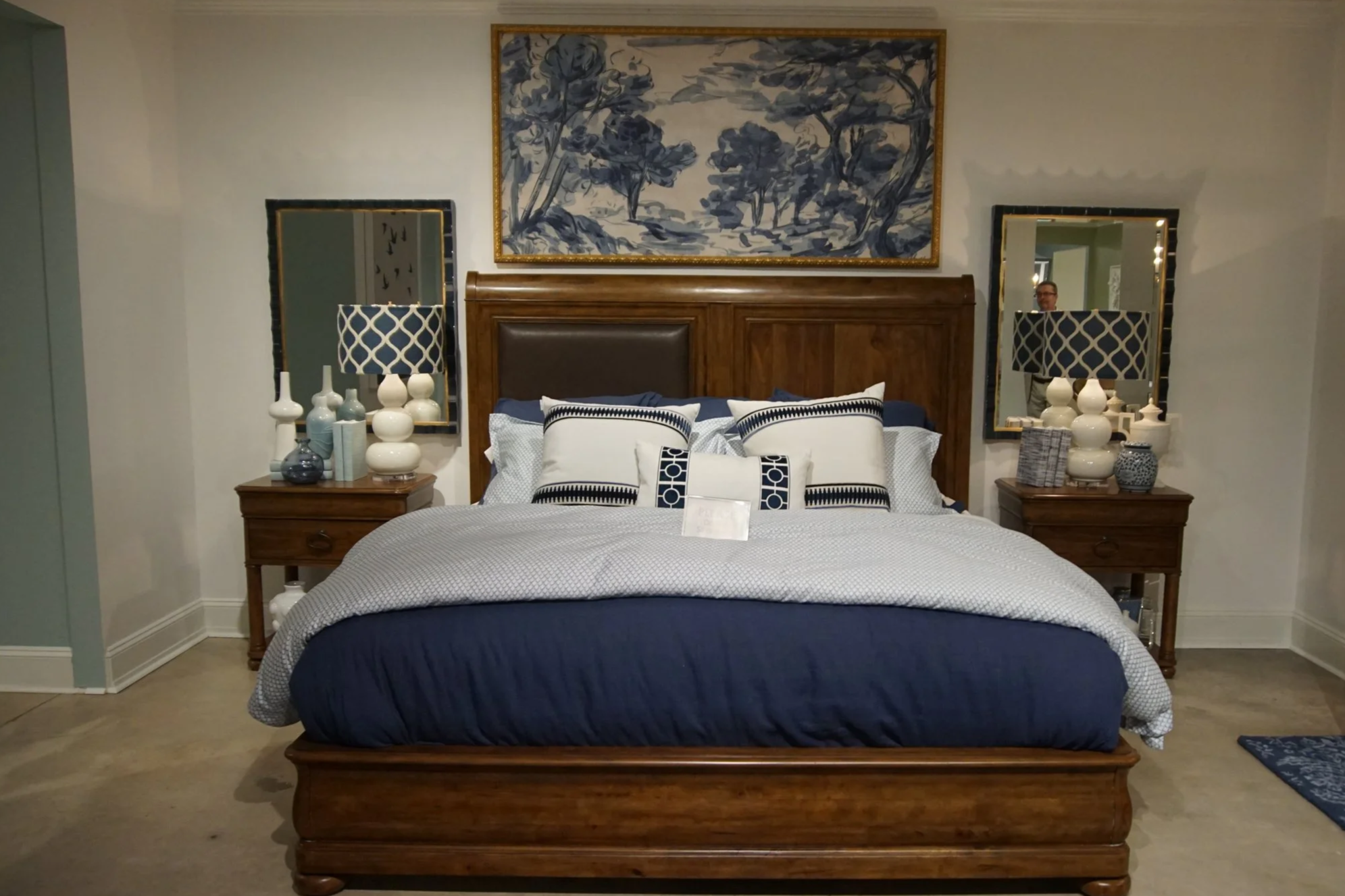A bedroom with a wooden bed frame and headboard, blue and white bedding, two nightstands with lamps and decorative vases, a large landscape-style painting above the bed, and mirrors on either side of the painting.
