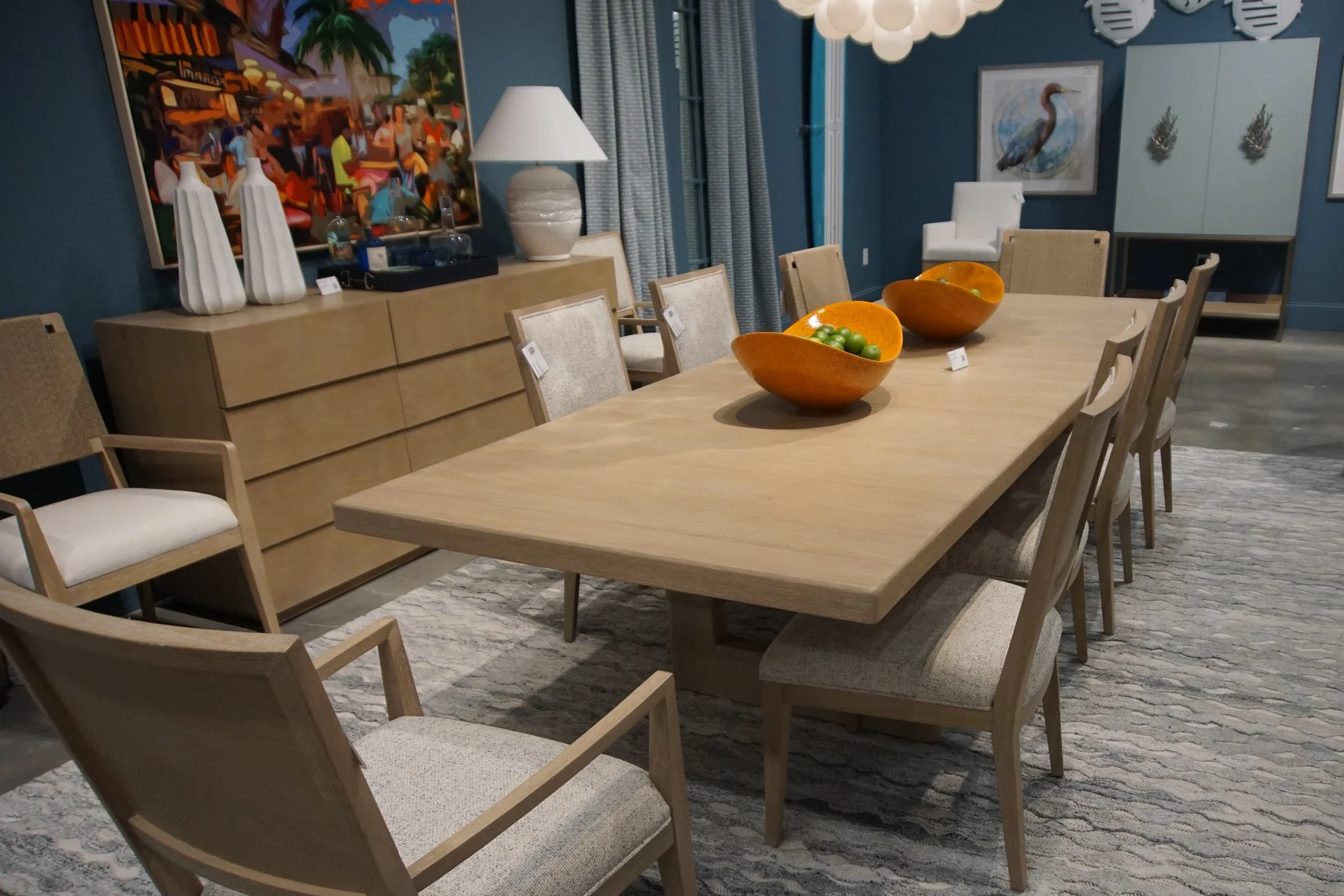 A dining room with a light wooden table and matching chairs, decorative bowls with green fruit, and various art pieces on the walls, including a colorful painting and animal portraits. Two people are in the room, one wearing a light gray plaid jacket