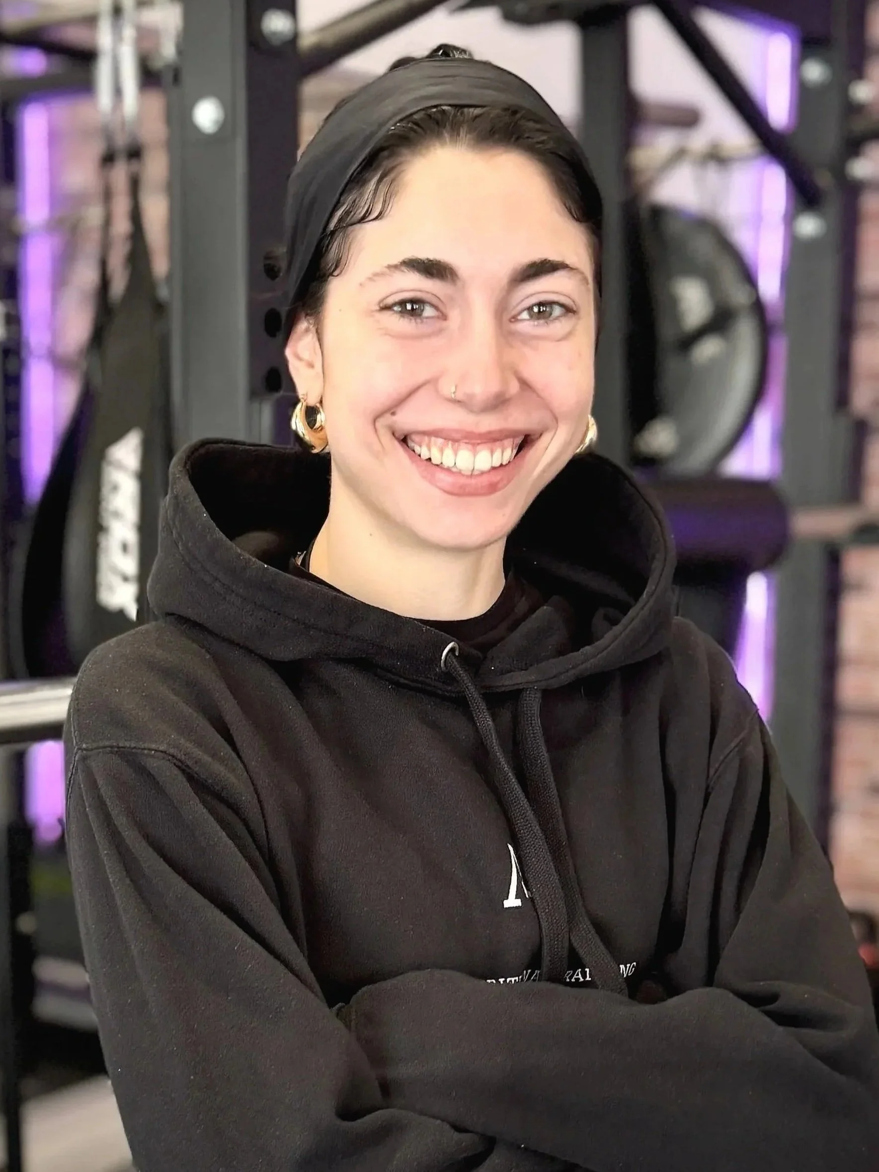 Junge Frau mit Brille und Dunkelhaar mit Zopf, die vor einer Hantelstange in einem Fitnessstudio steht, lächelnd in die Kamera schaut.