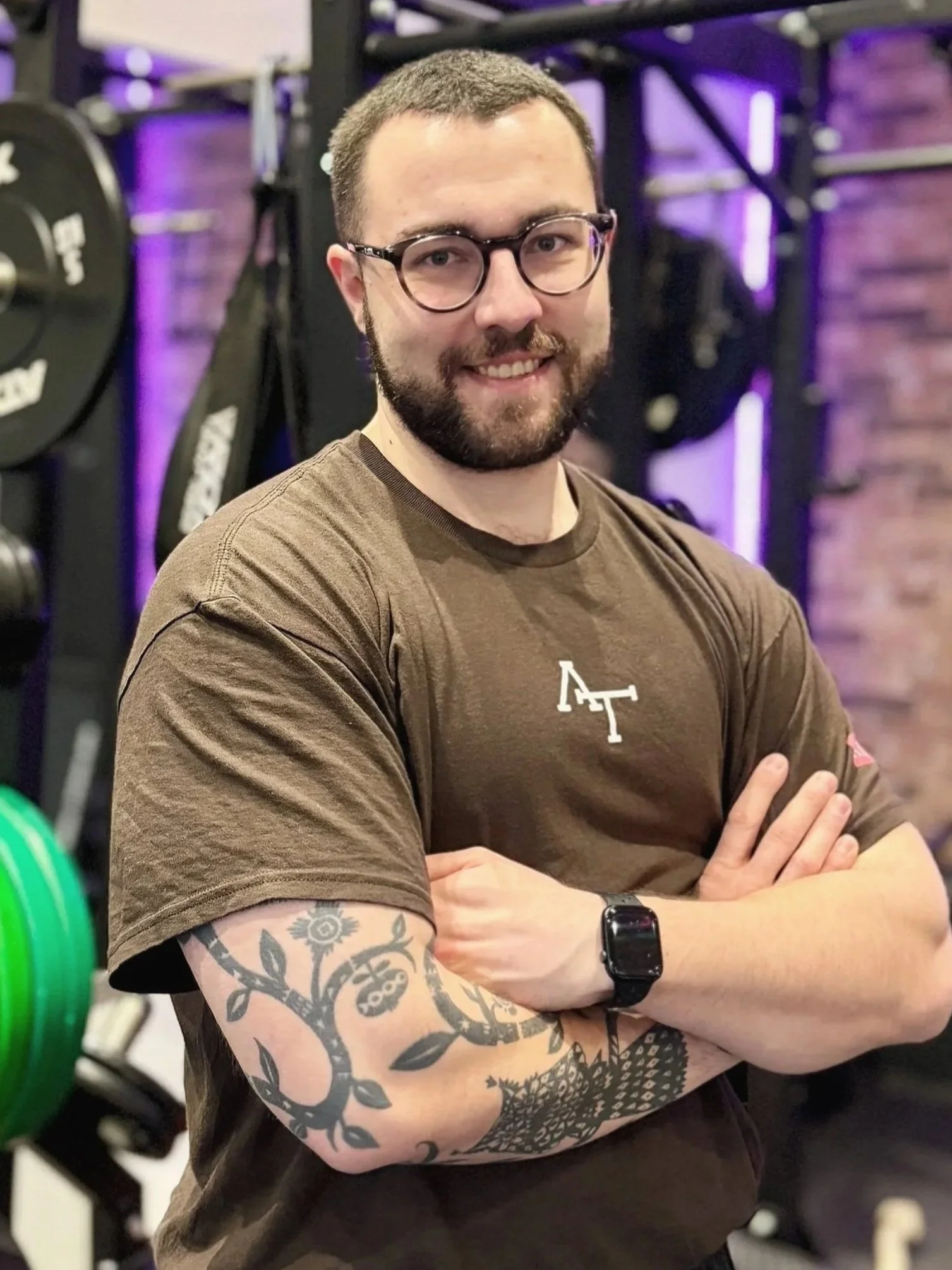 Ein Mann mit Tattoos, Brille und smartwatch, steht in einem Fitnessstudio vor einer Black-Box, umgeben von Hanteln, Gewichtsplatten und Kraftgeräten.