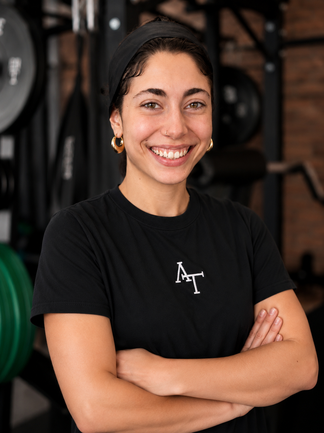 Junge Frau mit Brille und Dunkelhaar mit Zopf, die vor einer Hantelstange in einem Fitnessstudio steht, lächelnd in die Kamera schaut.