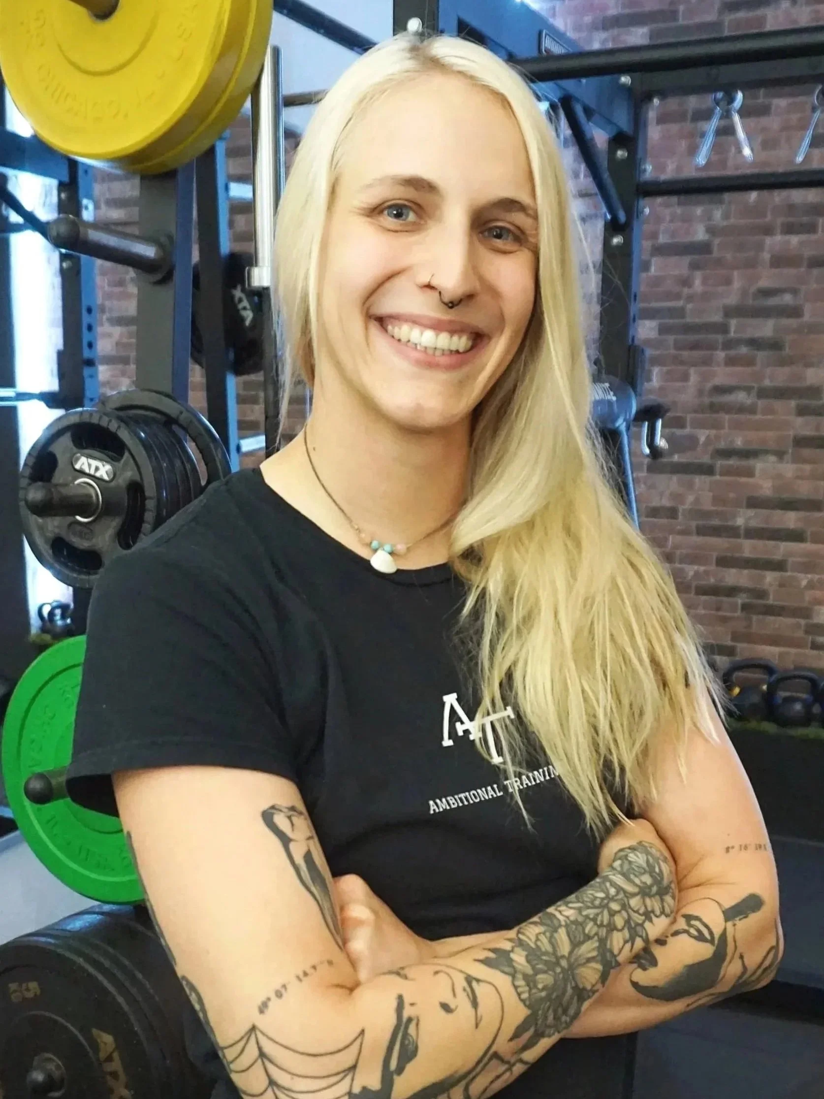 Junge Frau mit Tattoos, blonde Haare, lächelnd, in einer Fitnessstudio-Umgebung, trägt ein schwarzes T-Shirt und schwarze Shorts.
