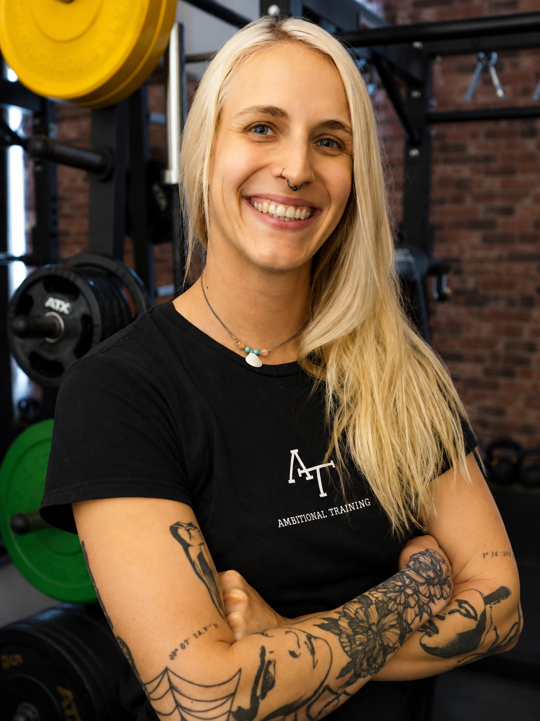 Junge Frau mit Tattoos, blonde Haare, lächelnd, in einer Fitnessstudio-Umgebung, trägt ein schwarzes T-Shirt und schwarze Shorts.