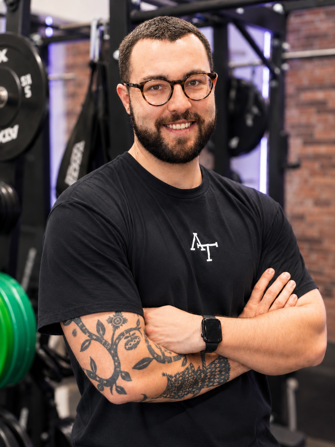 Ein Mann mit Tattoos, Brille und smartwatch, steht in einem Fitnessstudio vor einer Black-Box, umgeben von Hanteln, Gewichtsplatten und Kraftgeräten.
