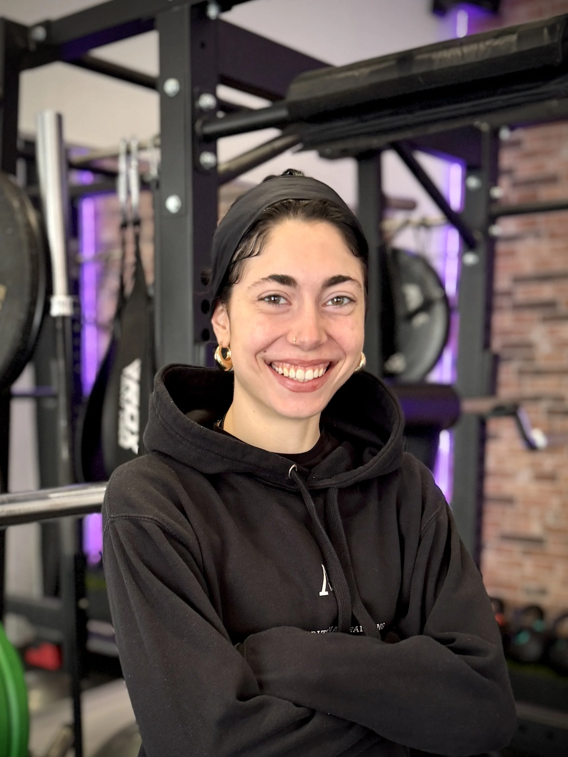 Junge Frau mit Brille und Dunkelhaar mit Zopf, die vor einer Hantelstange in einem Fitnessstudio steht, lächelnd in die Kamera schaut.