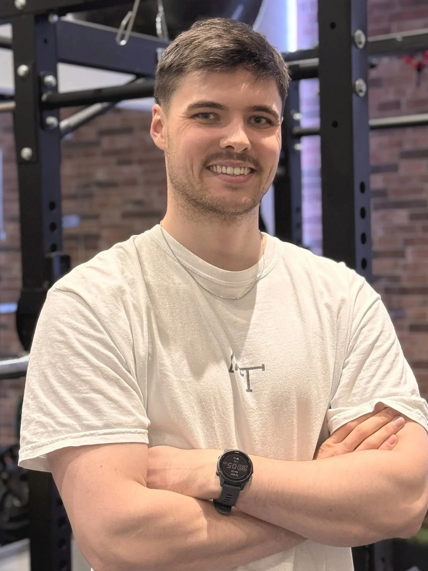 Ein junger Mann steht mit verschränkten Armen im Fitnessstudio vor einem schwarzen Kraftgerüst, trägt ein weißes T-Shirt und eine Armbanduhr, umgeben von Sportgeräten und einer Ziegelwand.