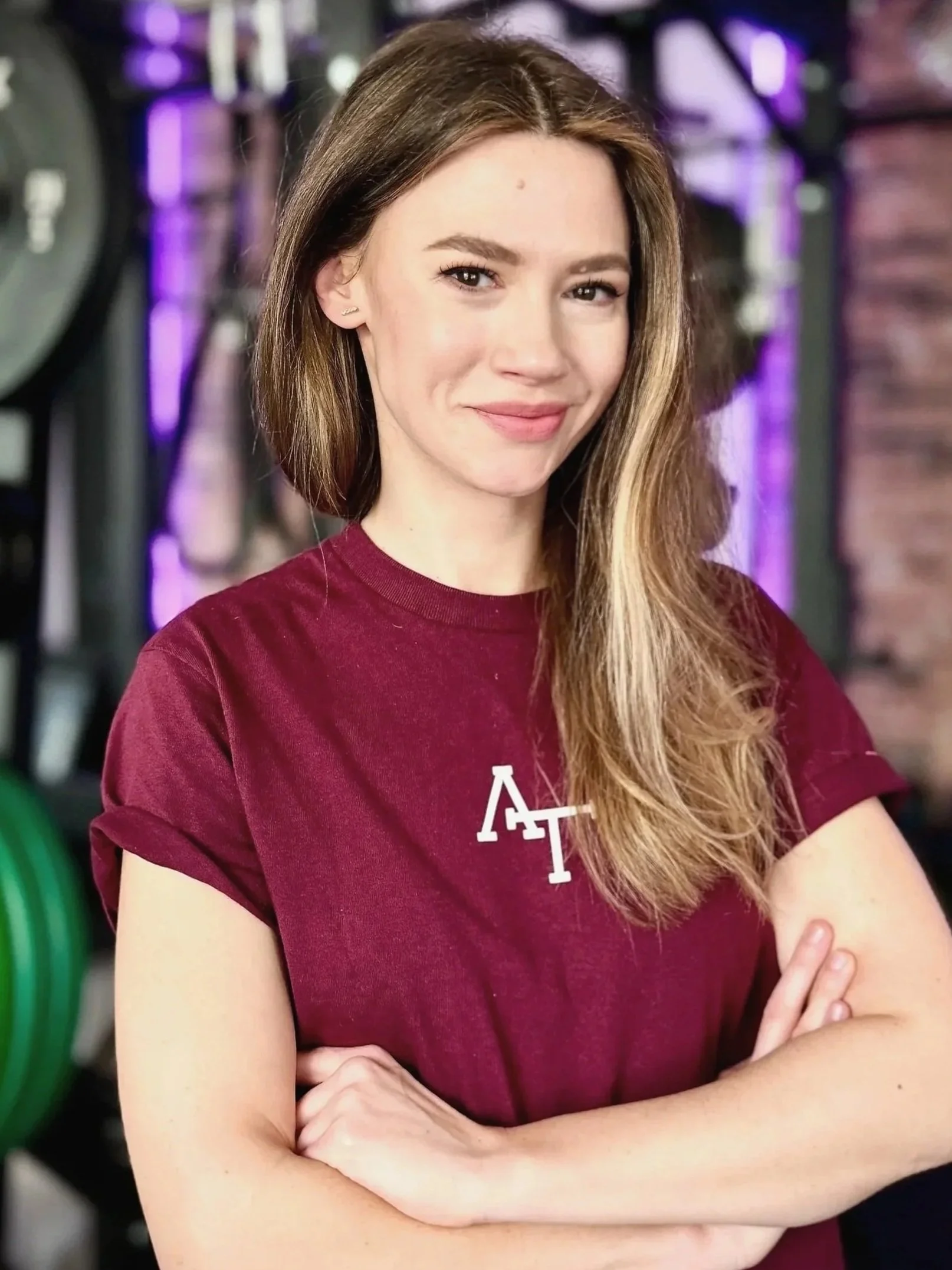 Junge Frau mit braunen Haaren in einem Fitnessstudio, lächelnd, trägt ein rotes T-Shirt mit weißen Buchstaben, vor einer Hantelbank gestanden.